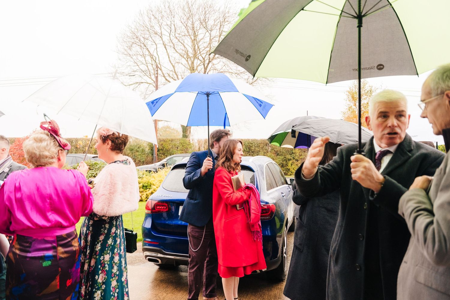 Wedding guests holding umbrellas while gathering outside in rainy weather.