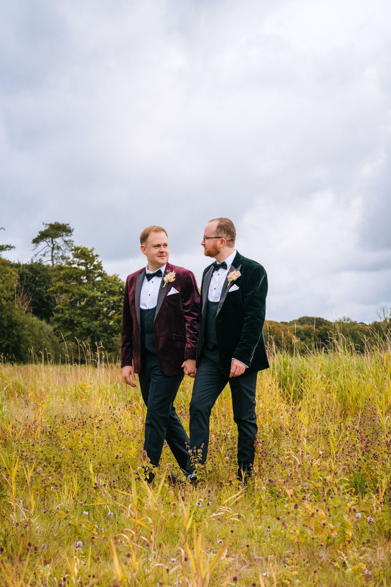 Two people in formal wear walking through a sunny meadow with tall grass and cloudy sky. LGBTQ Ashley Park Wedding Photographer