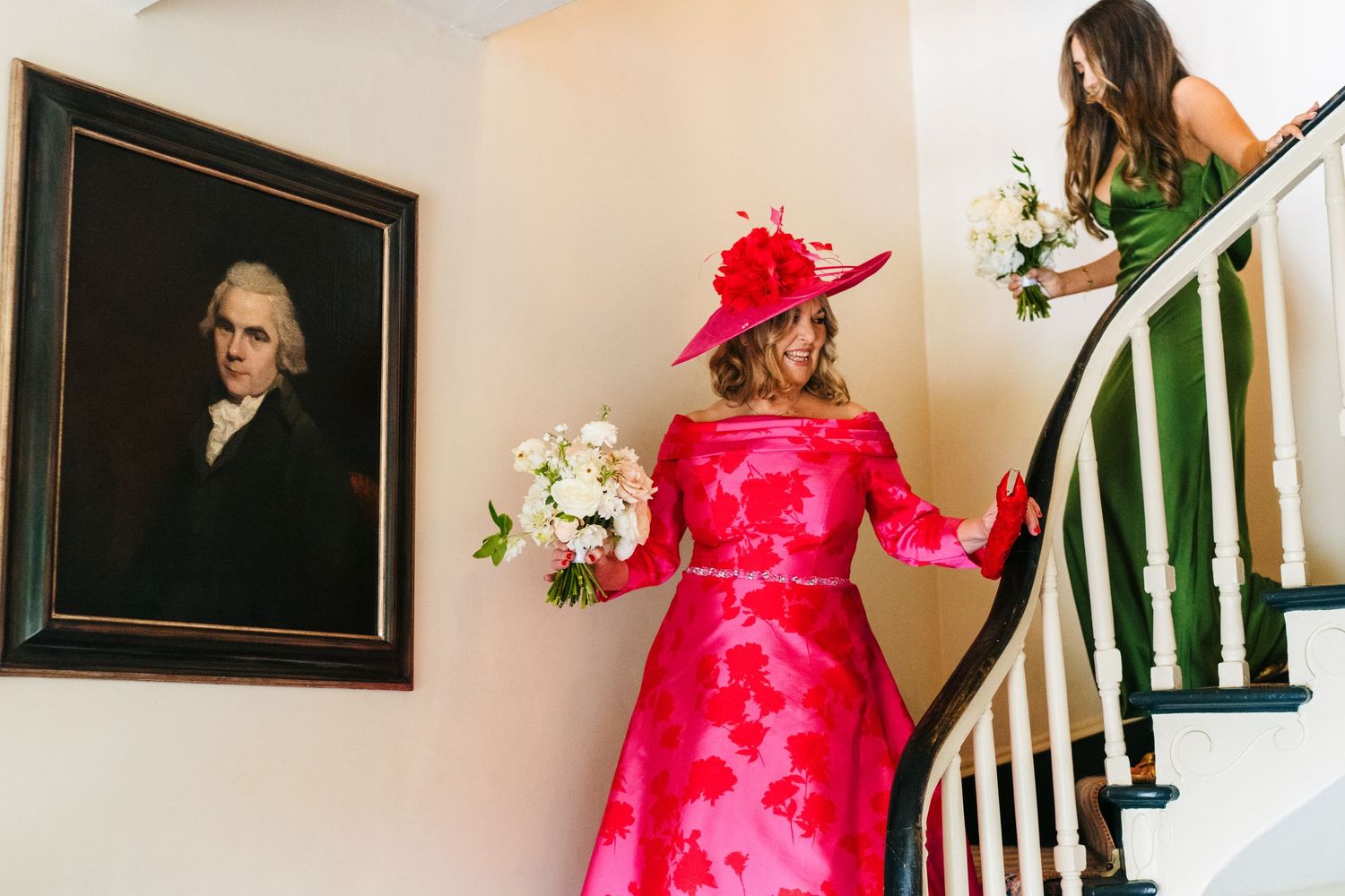 A woman in a vibrant red dress and hat descends a staircase with flowers near a formal portrait on the wall.  Virginia Park Lodge Wedding Ph