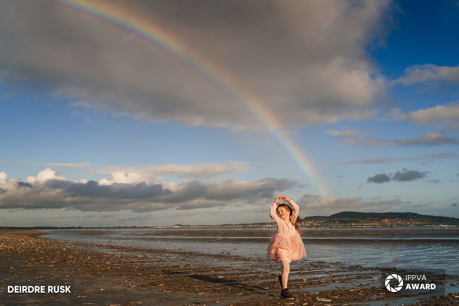 A small figure in pink twirls on a beach beneath a vibrant rainbow and dramatic cloudy sky. Family PHOTOGRAPHER Dublin PHOTOGRAPHER Dublin
