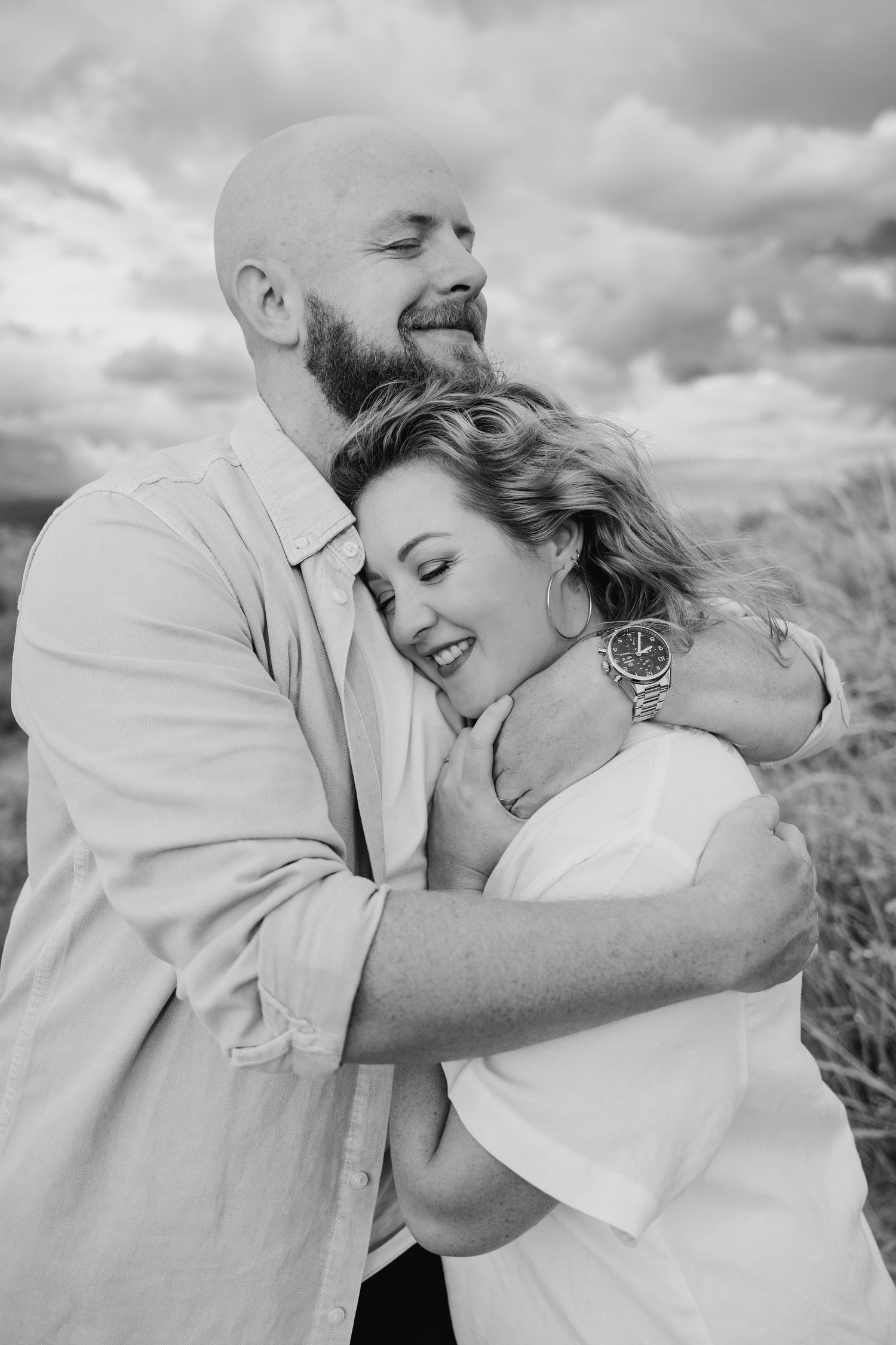 Black and white portrait of a couple embracing and laughing together outdoors with cloudy sky in background. PHOTOGRAPHER Dublin ireland