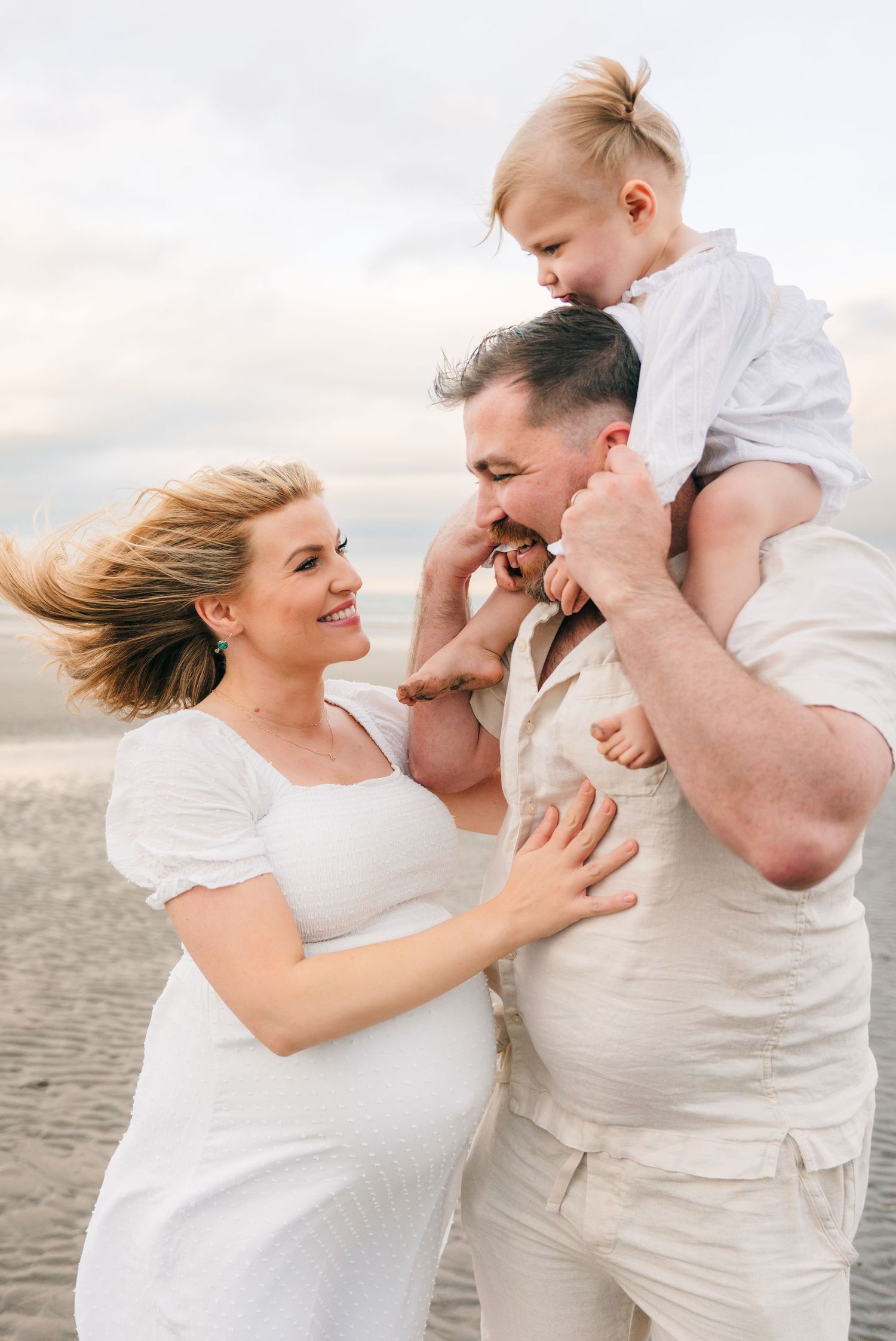 Family in white clothing sharing joyful moment together on beach at sunset with wind blowing through their hair. family PHOTOGRAPHER Dublin