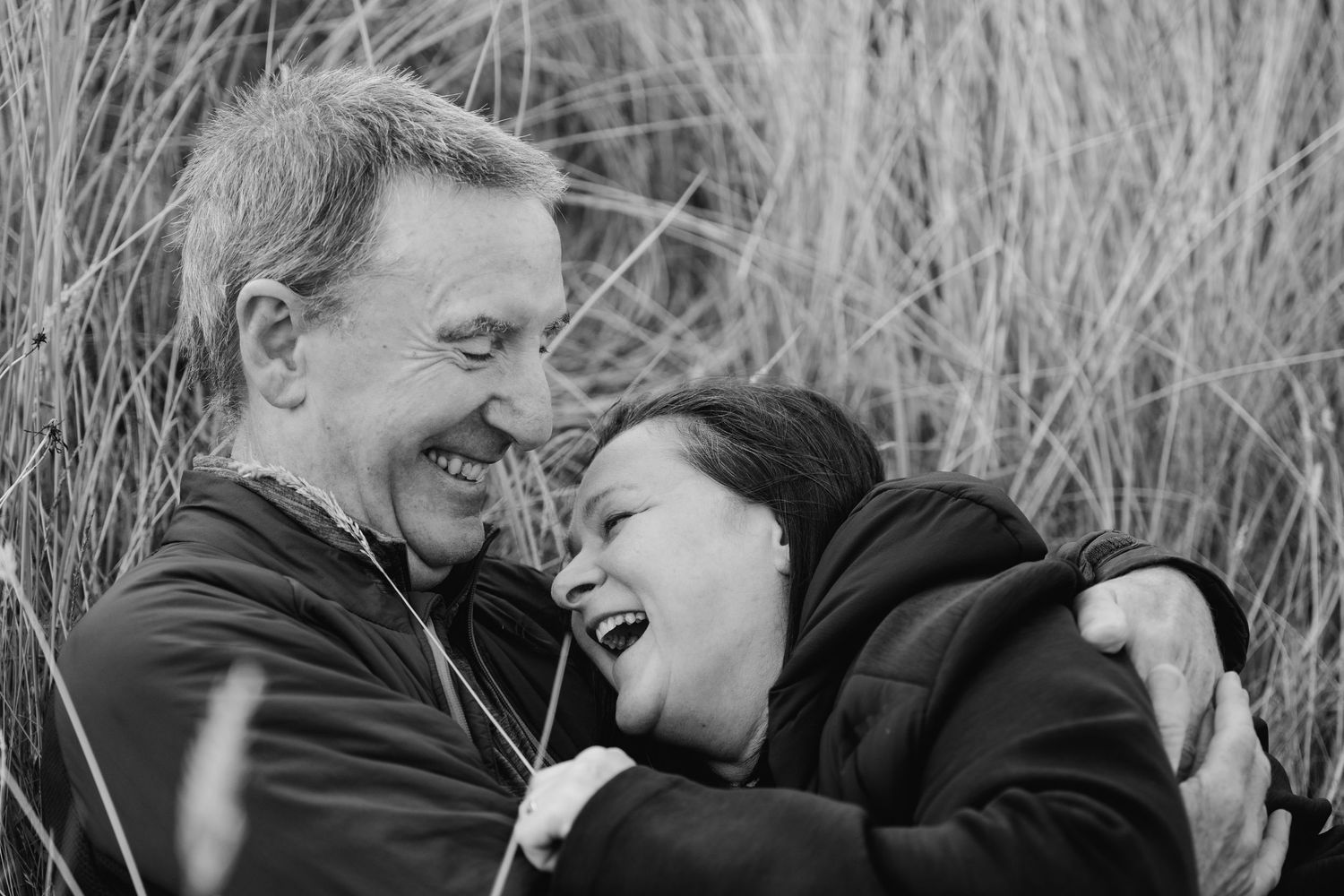 A candid black and white portrait shows a joyful moment of laughter between a couple amid tall grass. PHOTOGRAPHER Dublin ireland
