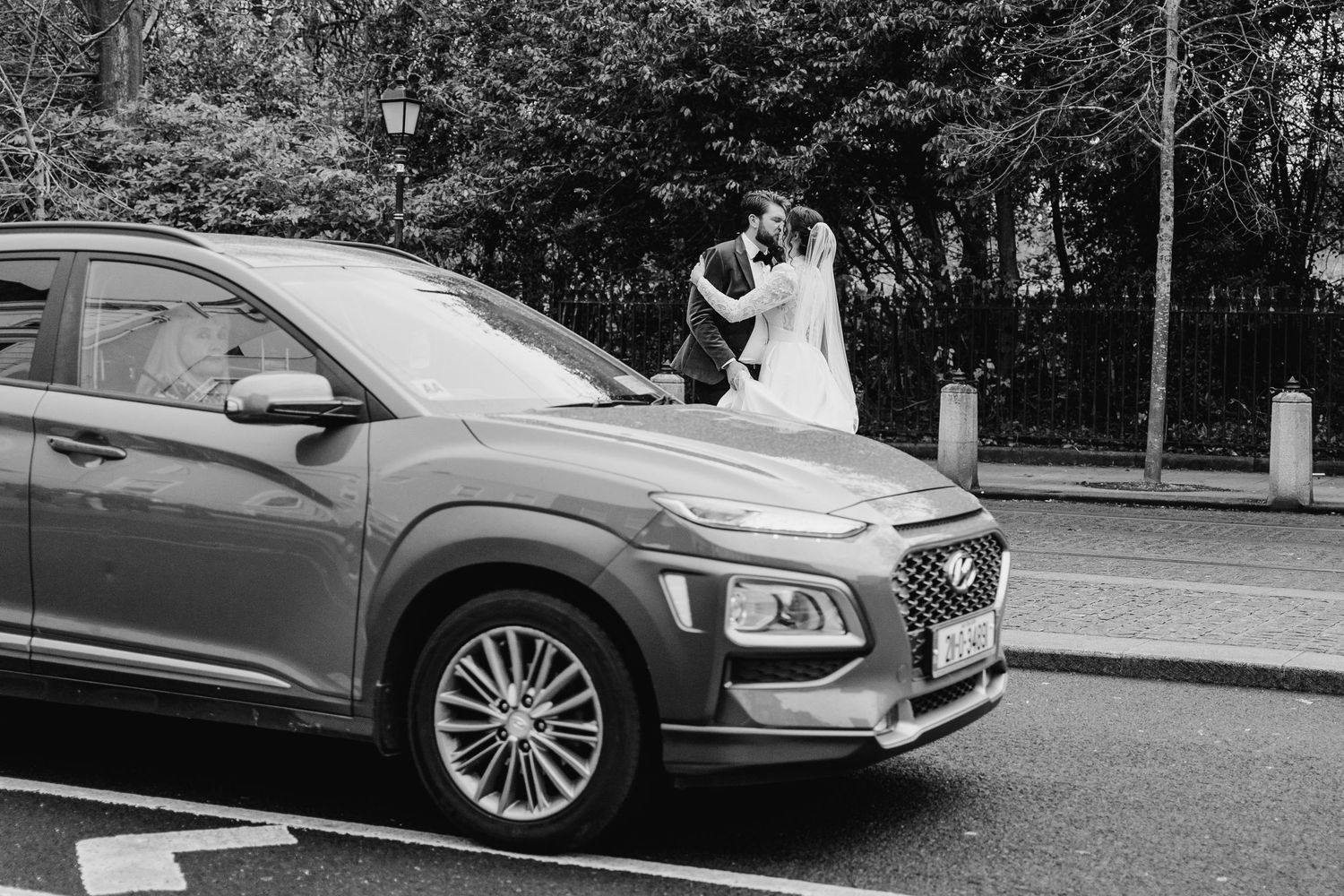 A couple in wedding attire stands beside a parked Hyundai Kona SUV in a black and white photograph. Stephens green club dublin Wedding photo