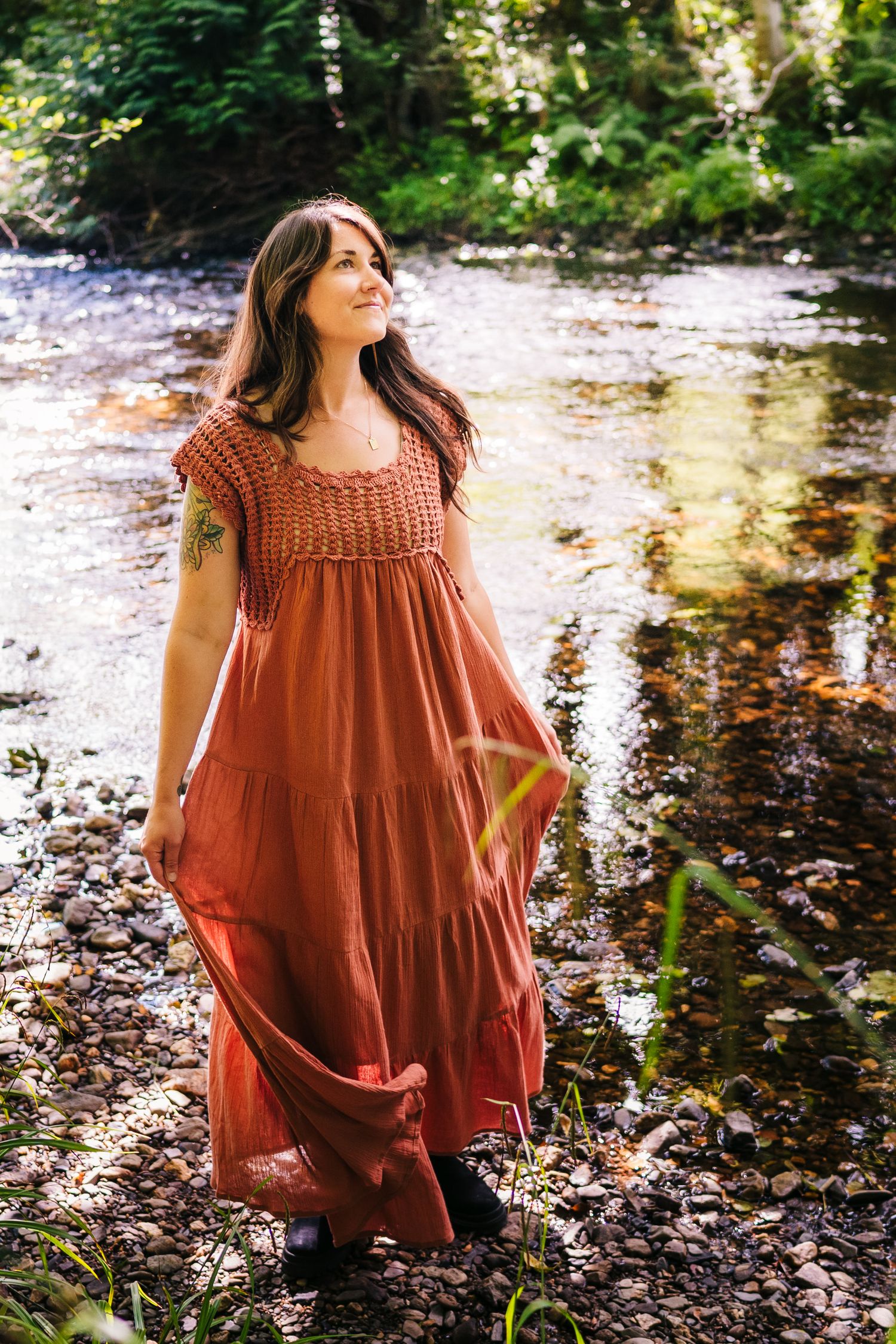 A rustic brown maxi dress with crochet details photographed by a stream in a woodland setting.