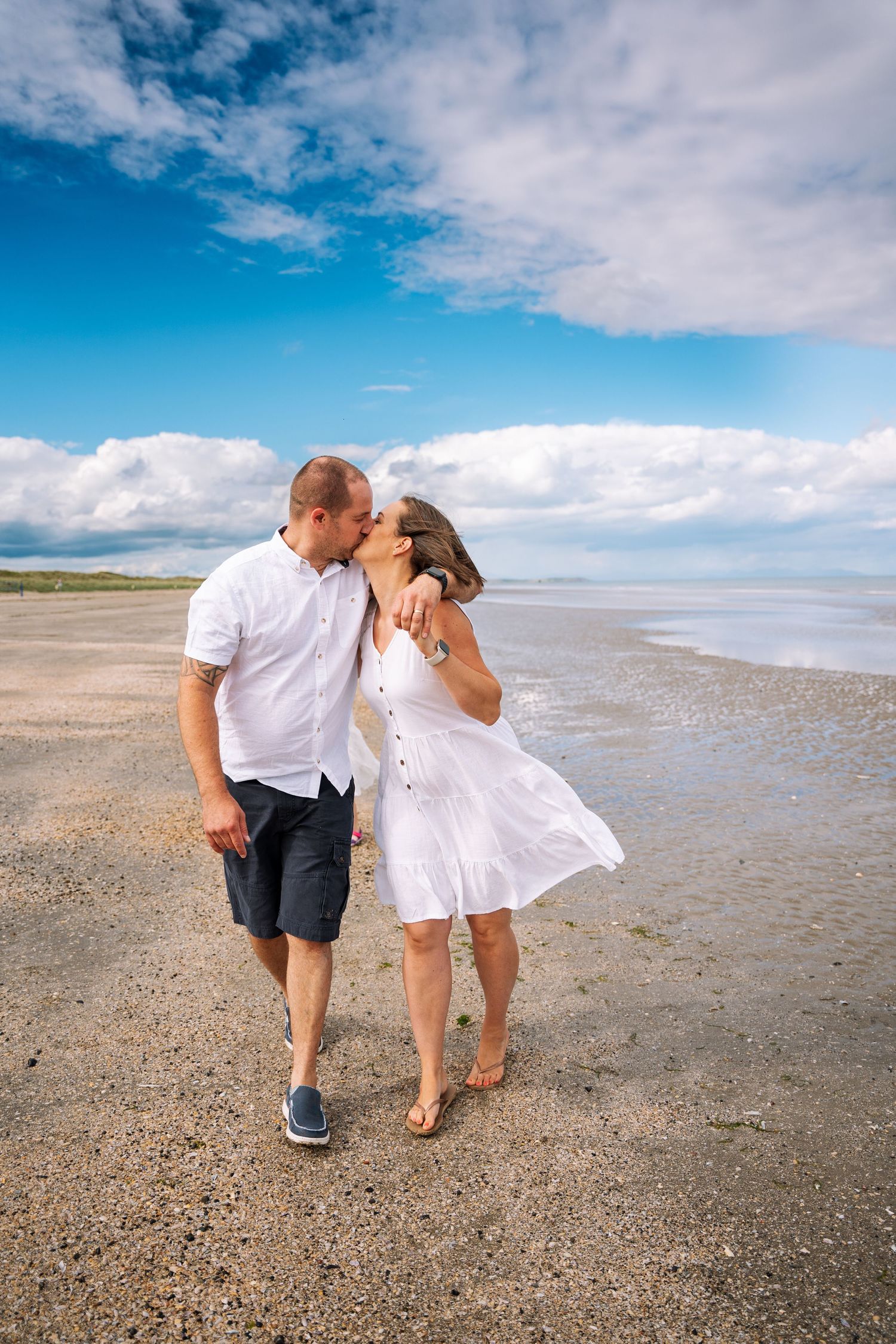 A couple shares a romantic moment on a sandy beach under blue skies with white fluffy clouds. PHOTOGRAPHER Dublin ireland