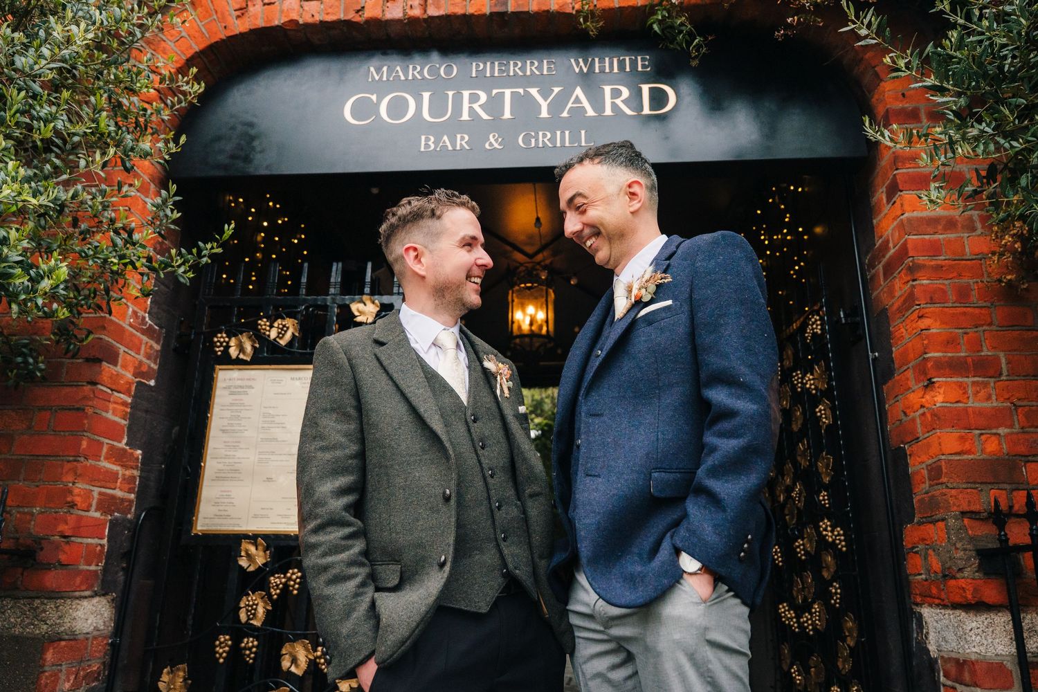 Wedding guests share a laugh in front of a courtyard entrance with brick facade and black awning.