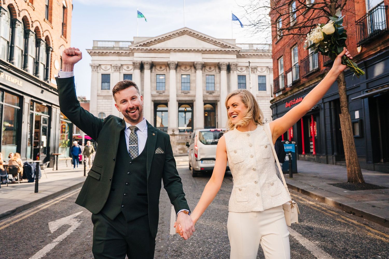 Couple celebrates with raised hands in front of historic city buildings and architecture. City Hall Dublin wedding photogrAPher