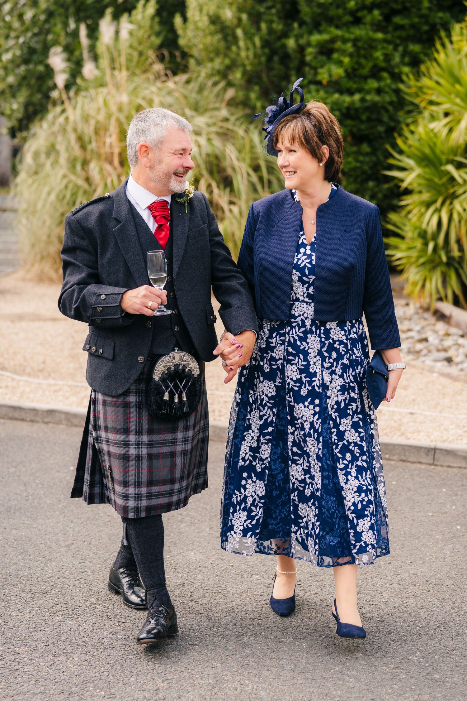 A couple in traditional Scottish wedding attire walks together, with the groom in a kilt and bride in a navy floral dress. Portmarnock Hotel