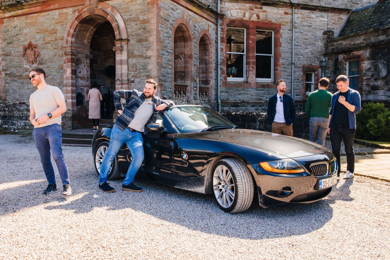A group stands around a sleek black sports car on a gravel driveway in front of a historic stone building. Castle Leslie Wedding Photographe