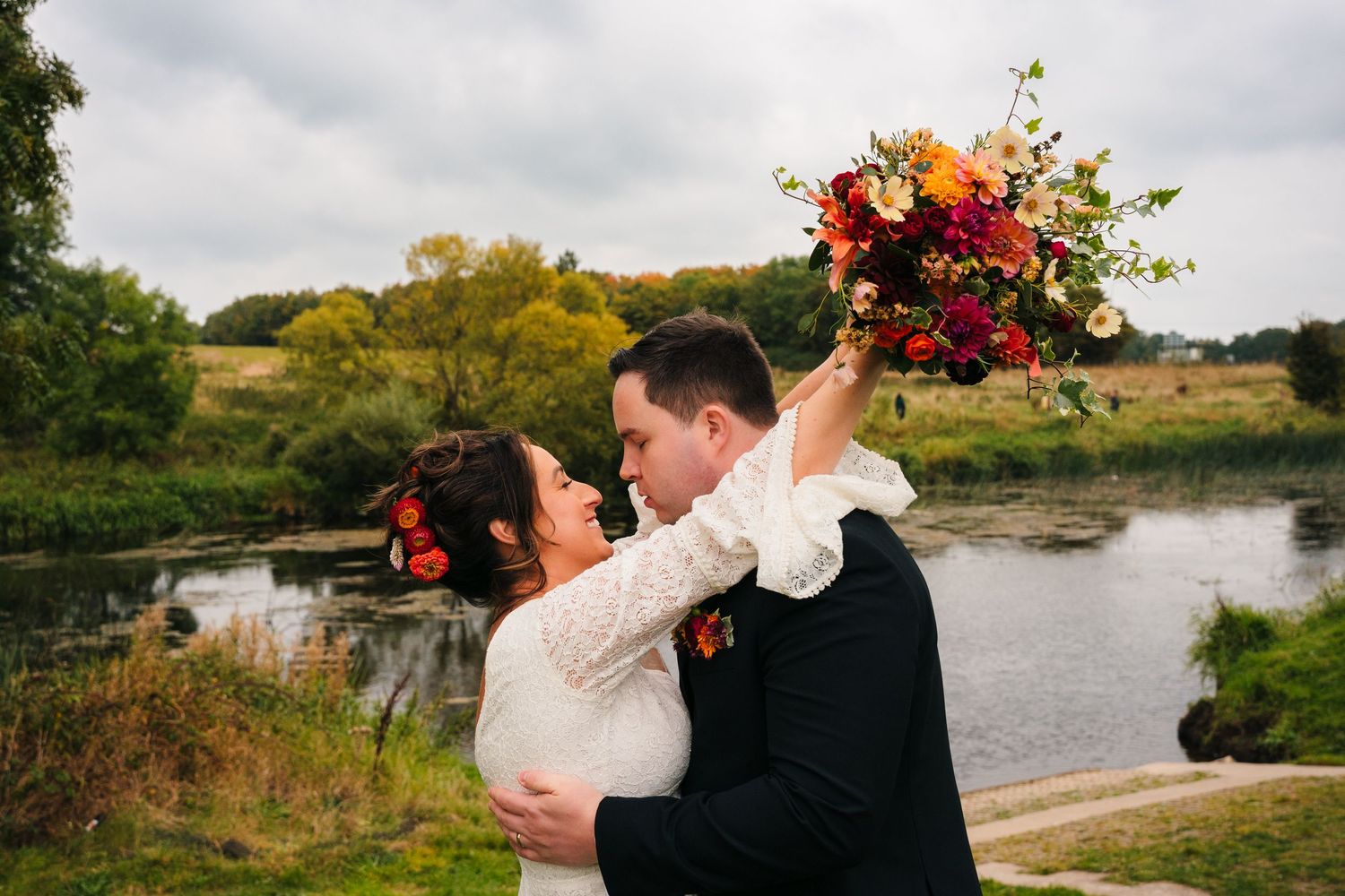 Bride and groom embrace by a pond with autumn foliage and vibrant orange bouquet.Wedding Photography Trim Castle Ireland