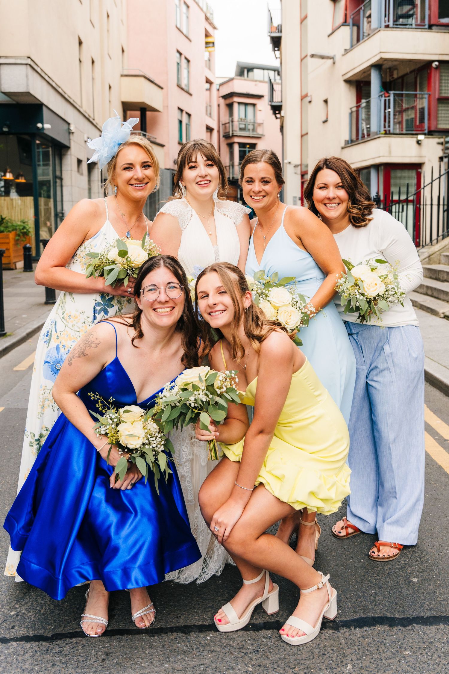 A group of bridesmaids in colorful dresses pose together on a city street holding white floral bouquets. PHOTOGRAPHER Dublin Ireland Wedding