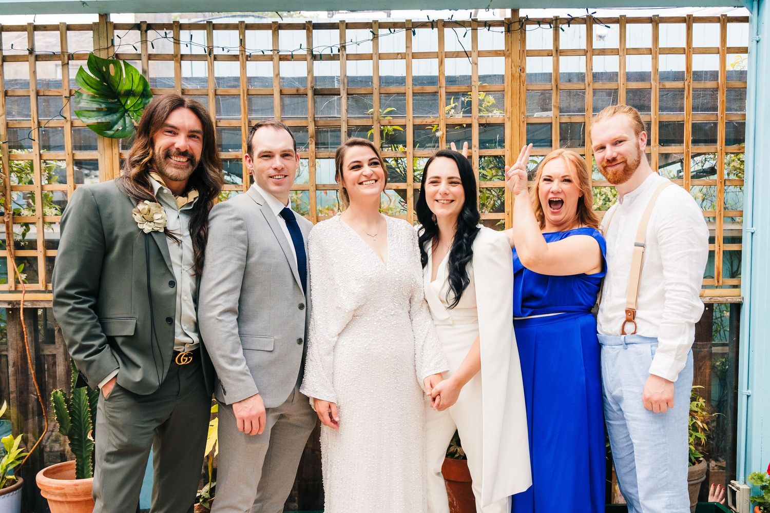 Wedding party poses together in formal attire against a wooden backdrop with greenery. House Dublin Wedding Photographer