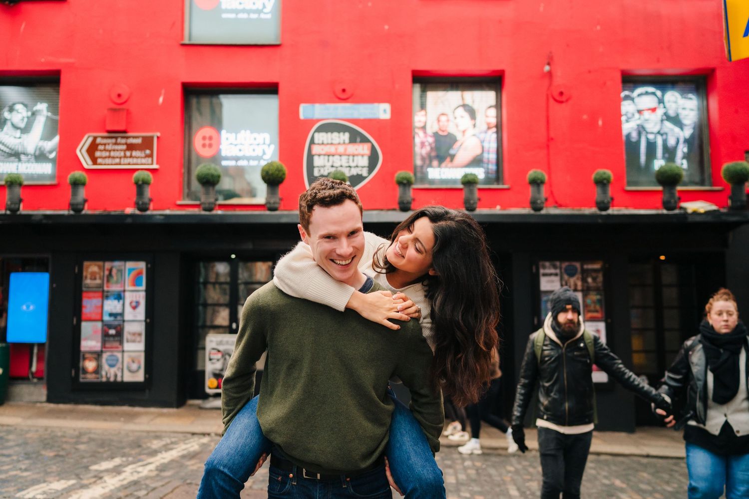 Couple having fun with piggyback ride in front of red building with vintage posters. Surprise proposal dublin photography