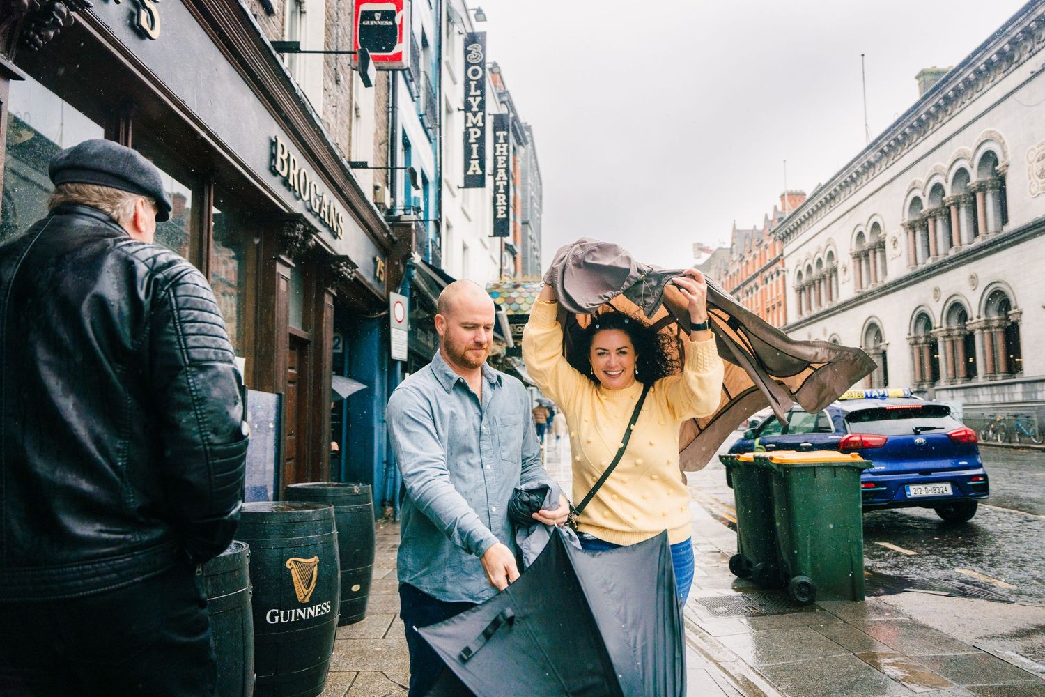 A couple shares a cheerful moment together while walking on a rainy day along a city street with historic buildings. Proposal Dublin Photogr