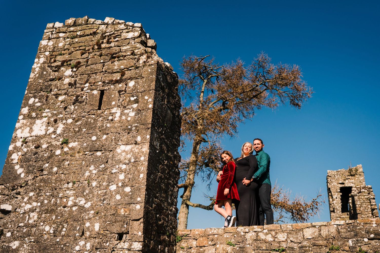 People stand between ancient stone castle towers against bright blue sky and autumn trees. Family PHOTOGRAPHER Dublin
