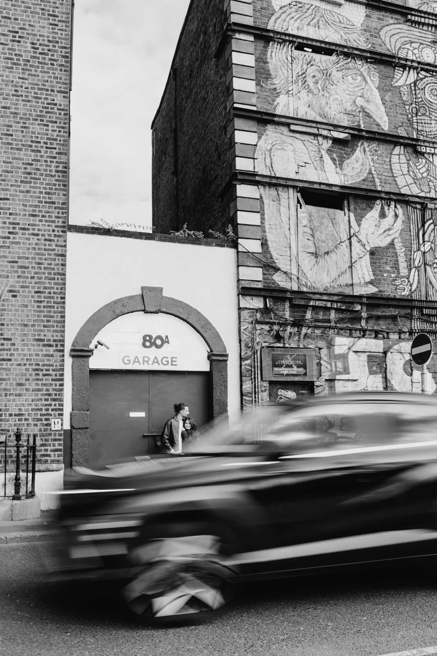 Black and white photo series of cars speeding past a brick building with '80s Garage' signage. Couple's Photoshoot PHOTOGRAPHER Dublin