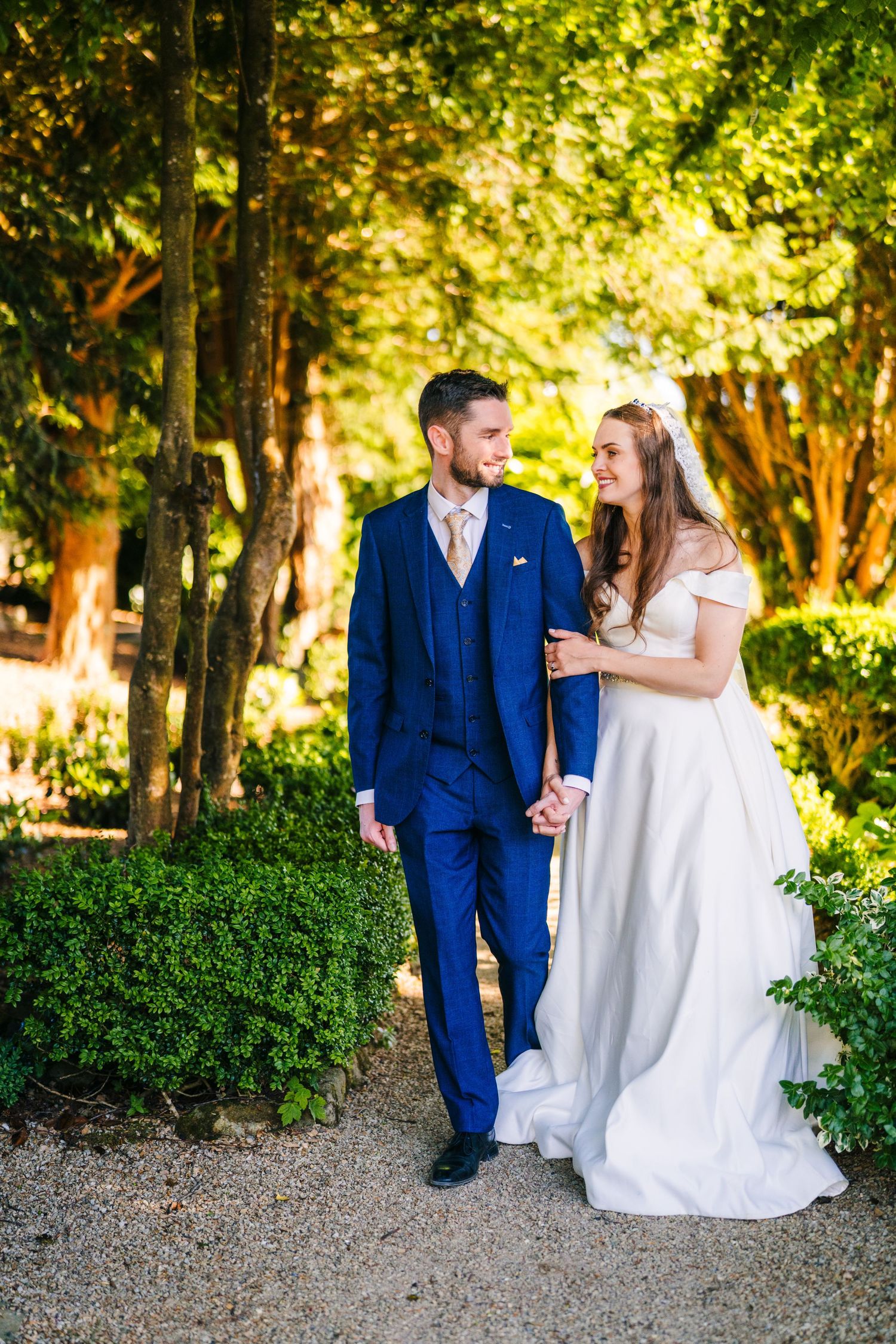 Poulaphouca Wedding photographA couple in wedding attire, with a navy suit and white gown, share a tender moment in a sunlit garden setting.