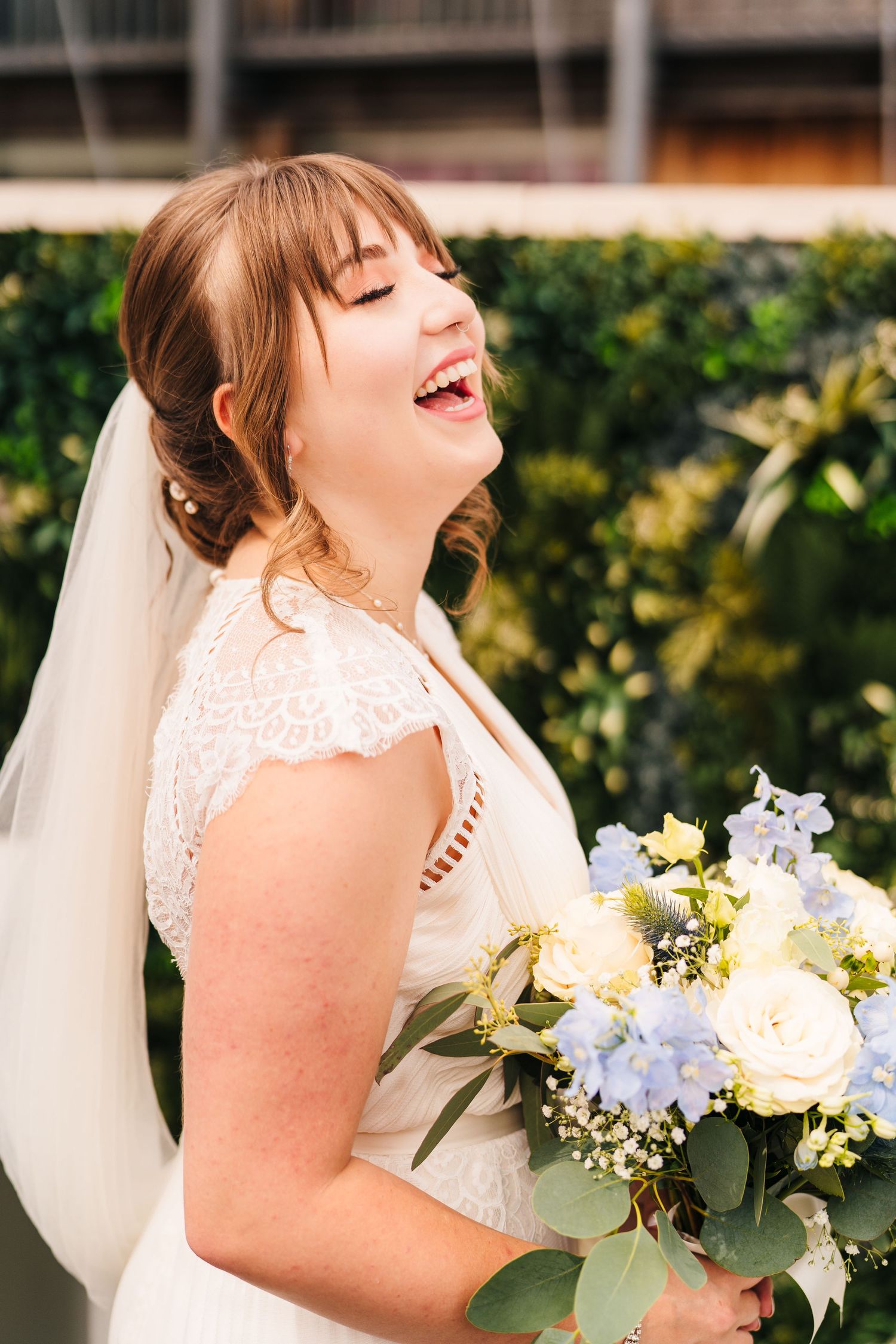 A joyful bride in a lace dress laughs while holding a bouquet of white and blue flowers with greenery. PHOTOGRAPHER Dublin Ireland Wedding