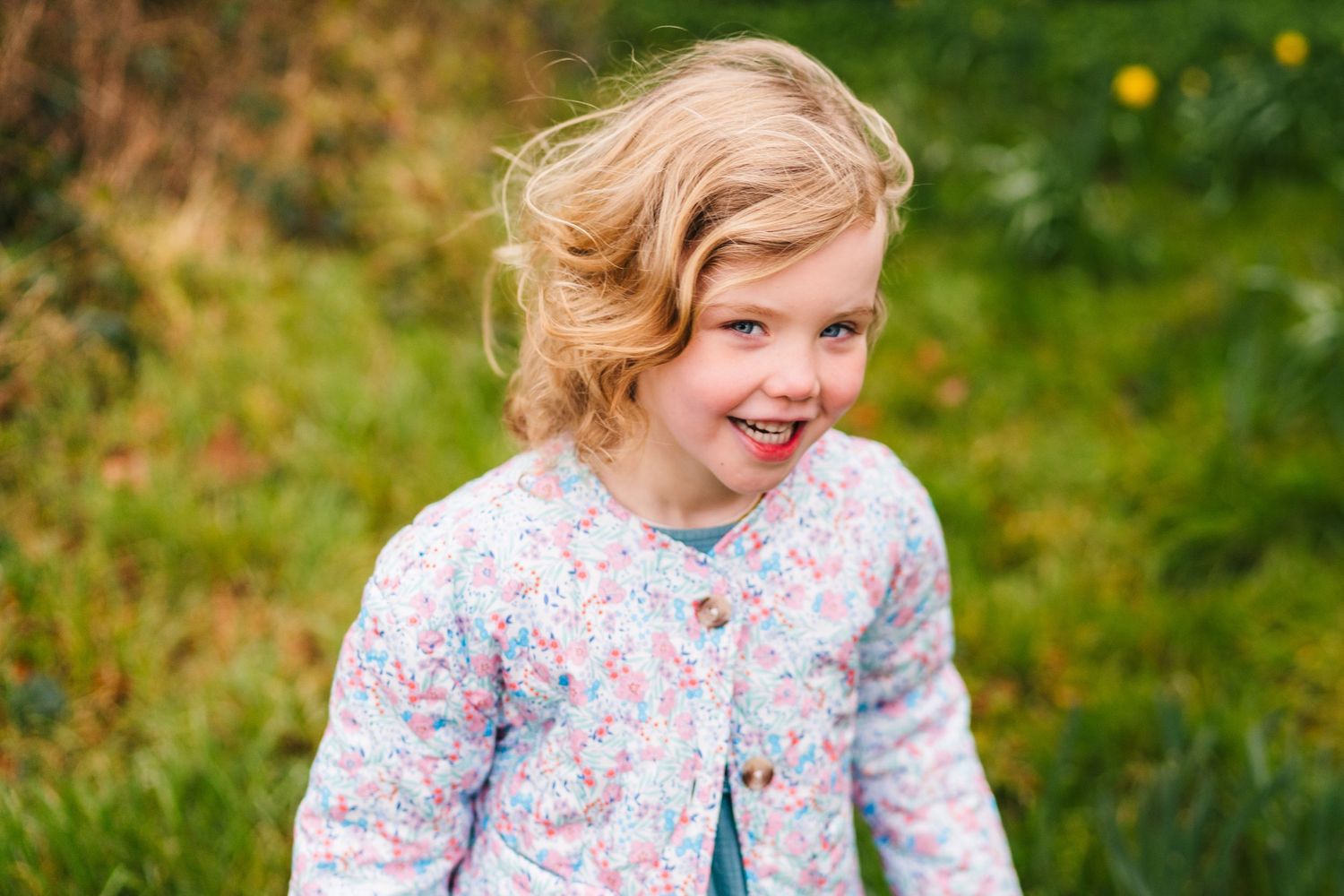 Family photographer DublinChild wearing a floral print cardigan smiles joyfully while playing outdoors in a grassy area with yellow flowers.