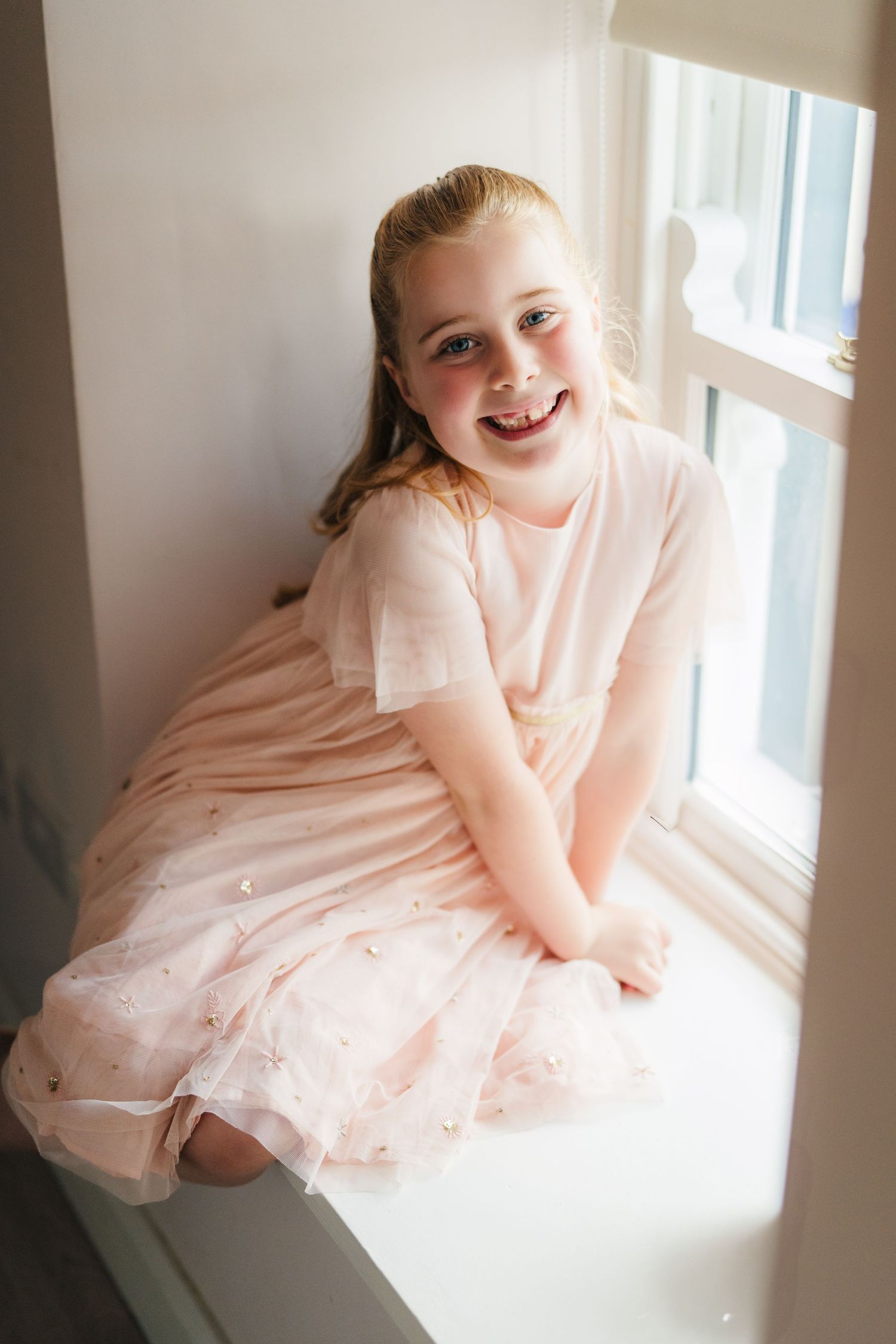 A young child in a pale pink tulle dress sits by a window with natural light streaming in. . Village Barn Tyrrellspass wedding photographer