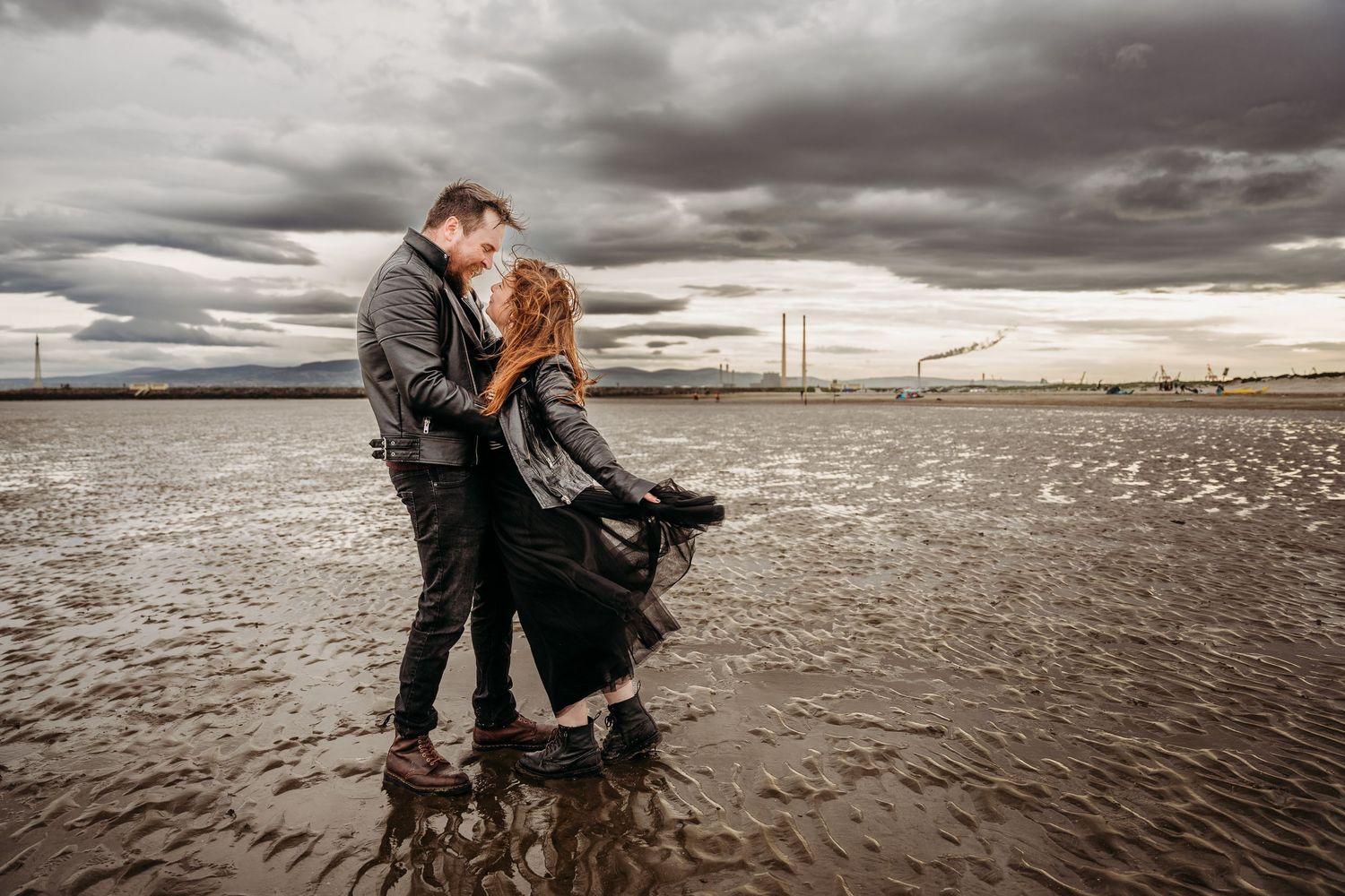 A couple embraces on a moody beach with dramatic storm clouds overhead and waves breaking on the shore. engagement PHOTOGRAPHER Dublin