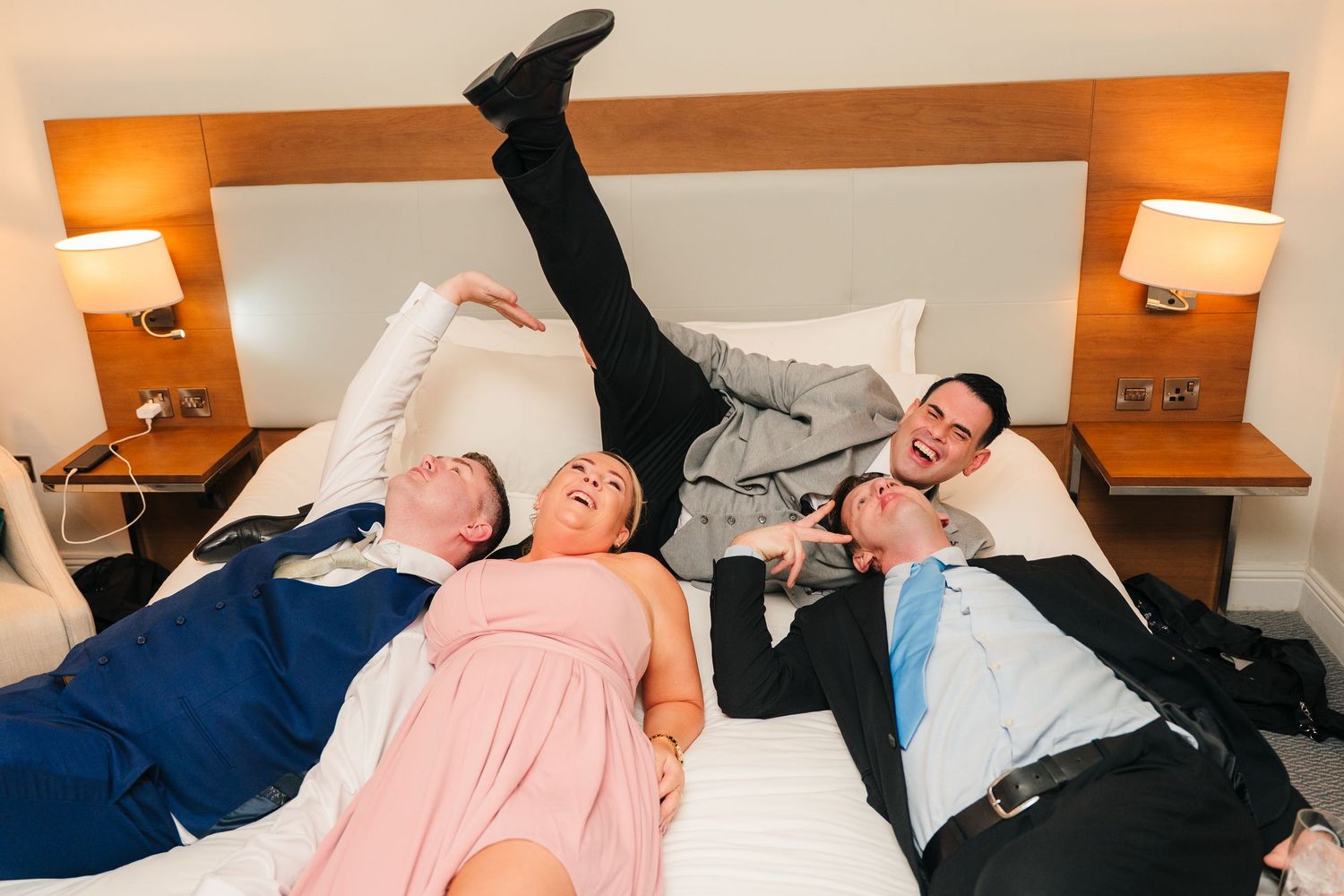 Wedding party members laugh while laying together on a hotel bed in formal attire. Portmarnock Hotel Wedding PHOTOGRAPHER Dublin