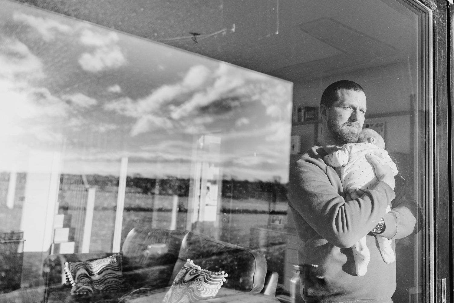 Black and white photo of reflection in window showing someone holding a baby against cloudy sky. Family PHOTOGRAPHER Dublin