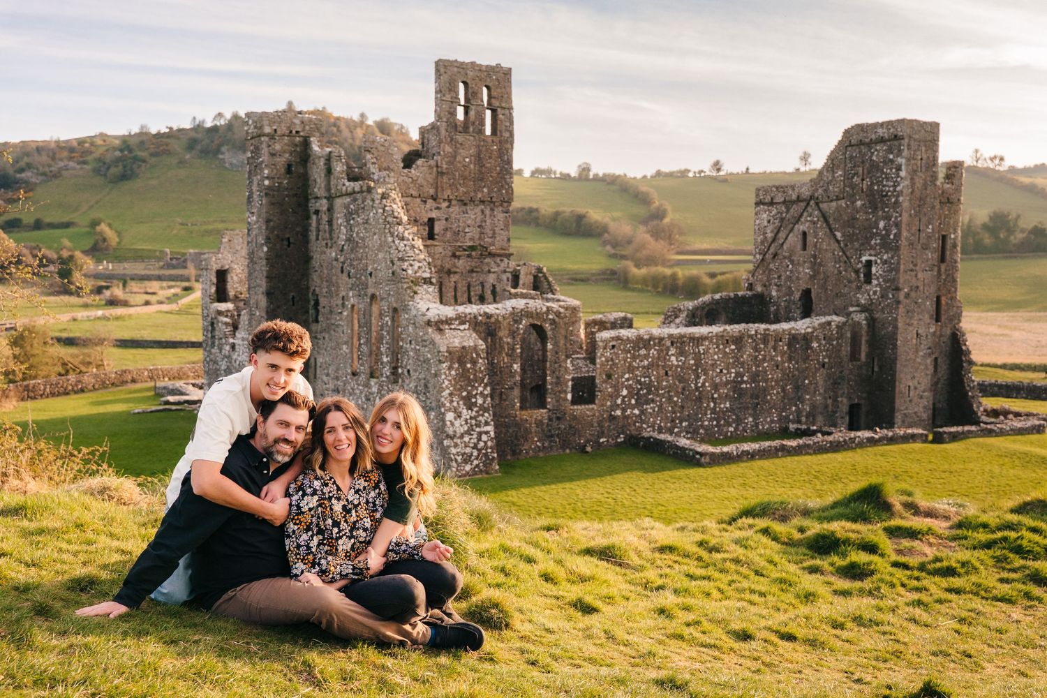 Couple sitting together on grass near historic stone castle ruins during sunset engagement photoshoot. Photographer Ireland dublin