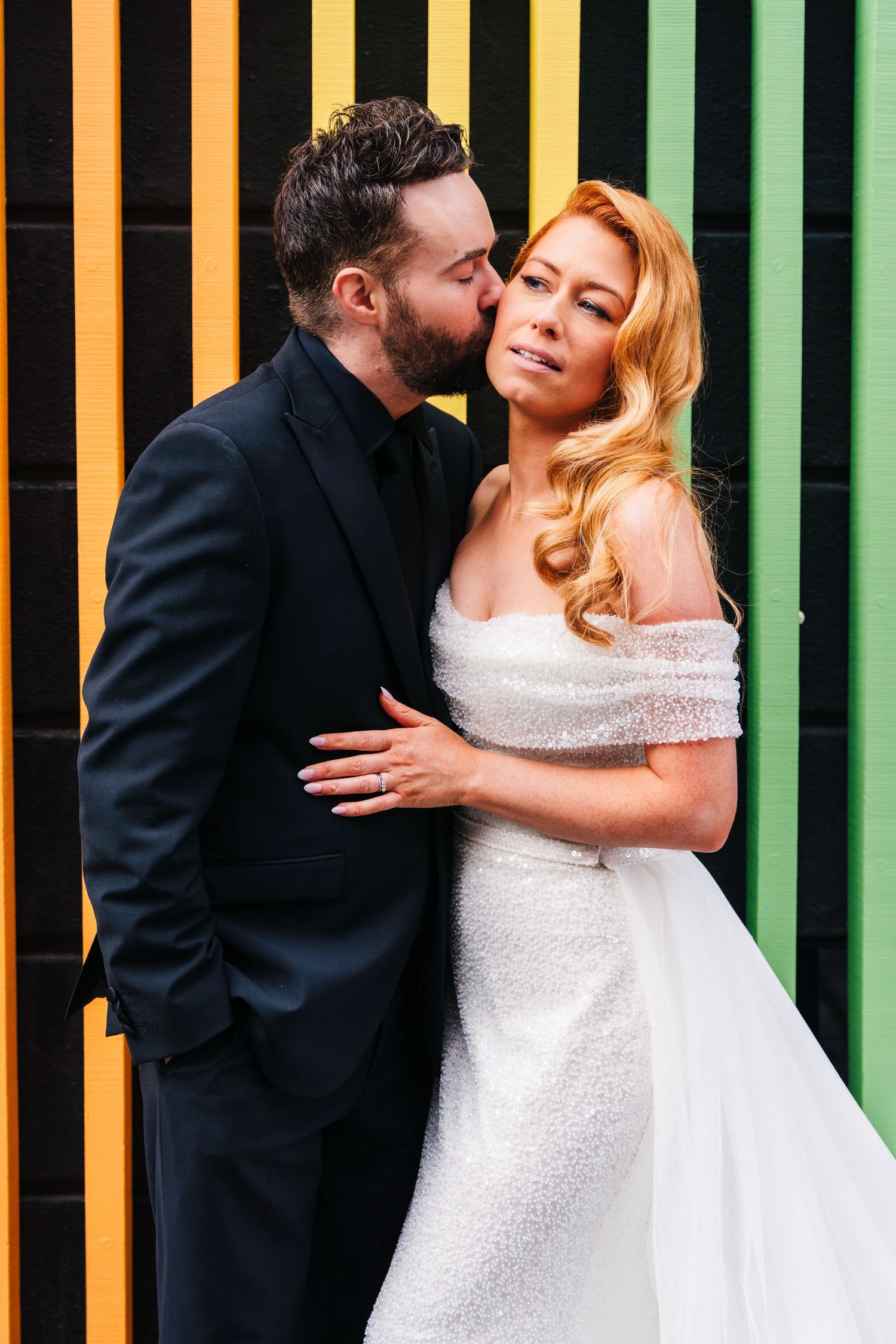 A bride in a white off-shoulder gown and groom in black suit embrace against a colorful striped wall.
