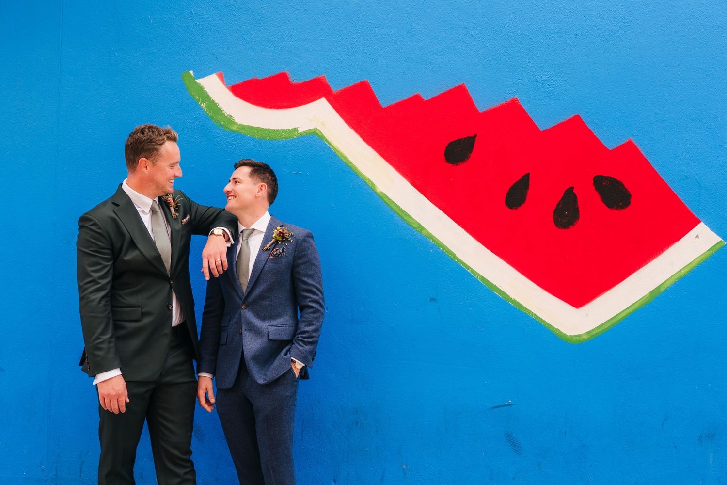 Two people in suits stand in front of a vibrant blue wall with a large painted watermelon slice artwork.