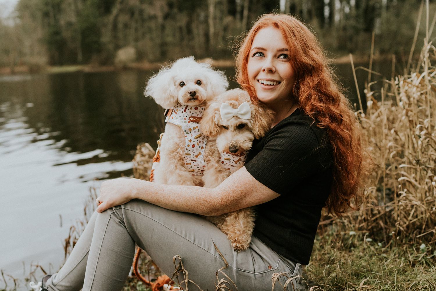 A woman with flowing red hair sits by a lake holding two small fluffy white dogs among tall grass at golden hour. PHOTOGRAPHER Dublin irelan
