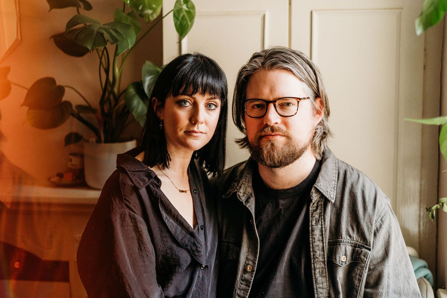 Couple sitting together with black bangs and glasses wearing dark clothing against white wall with leafy plants. PHOTOGRAPHER Dublin ireland