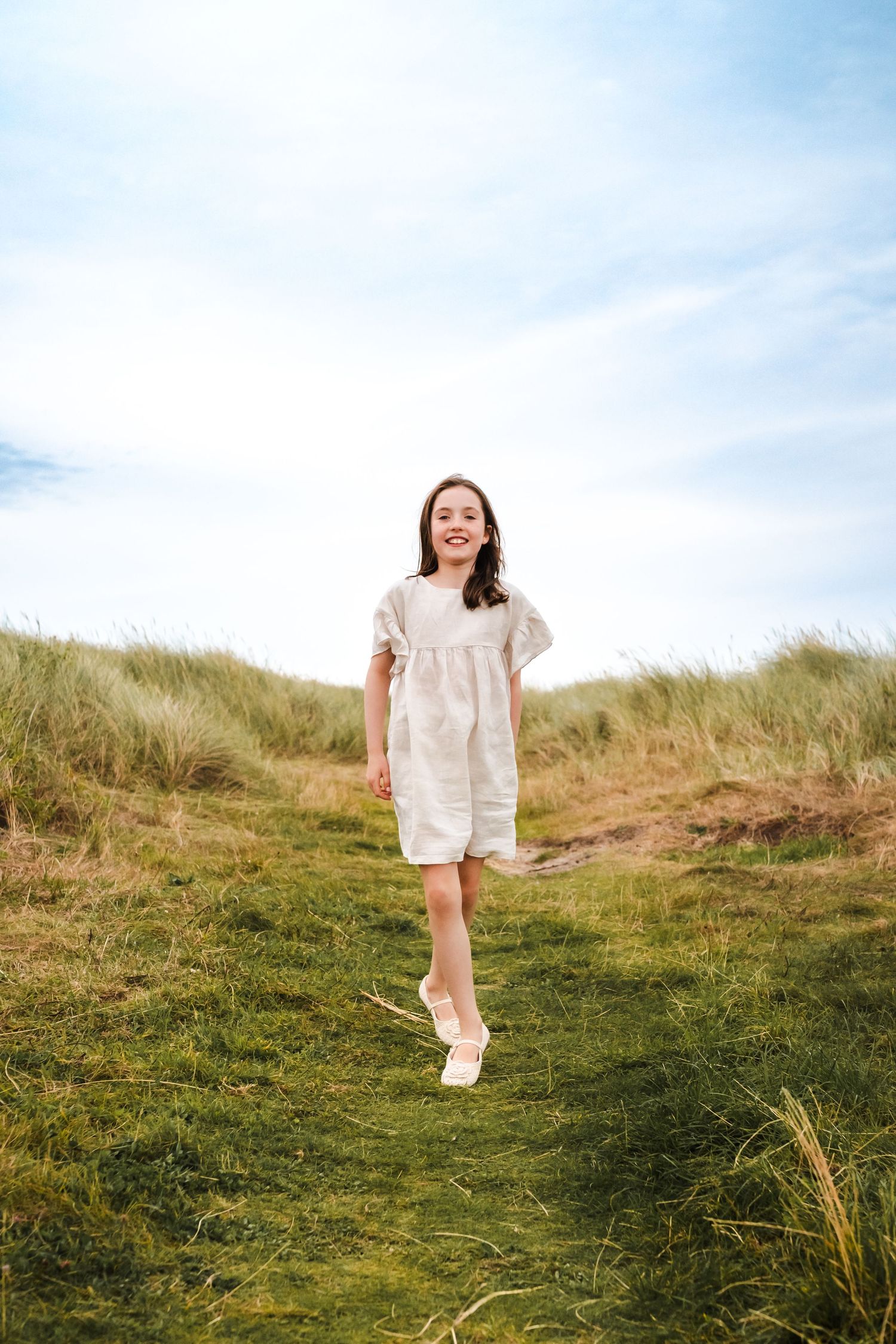 Person in a white dress walks along a grassy coastal hillside under blue skies.  Family photographer Dublin Ireland