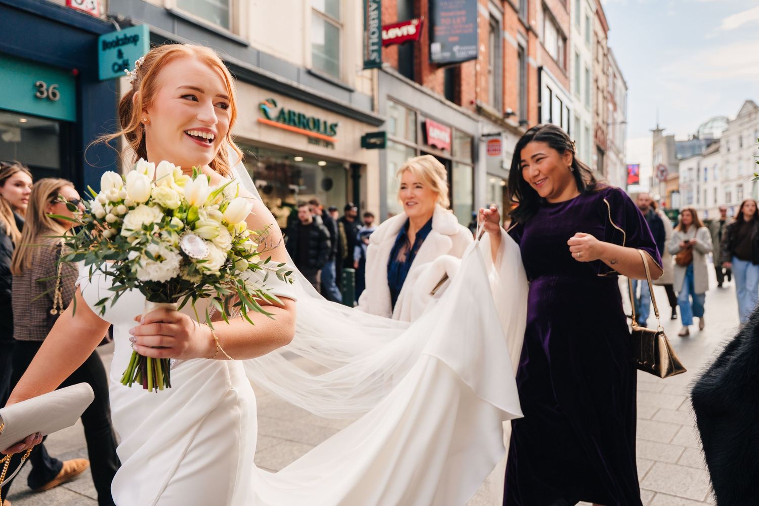 A bride in a white dress holds a bouquet while walking down a busy city street with her wedding party. Wedding Photographer Dublin