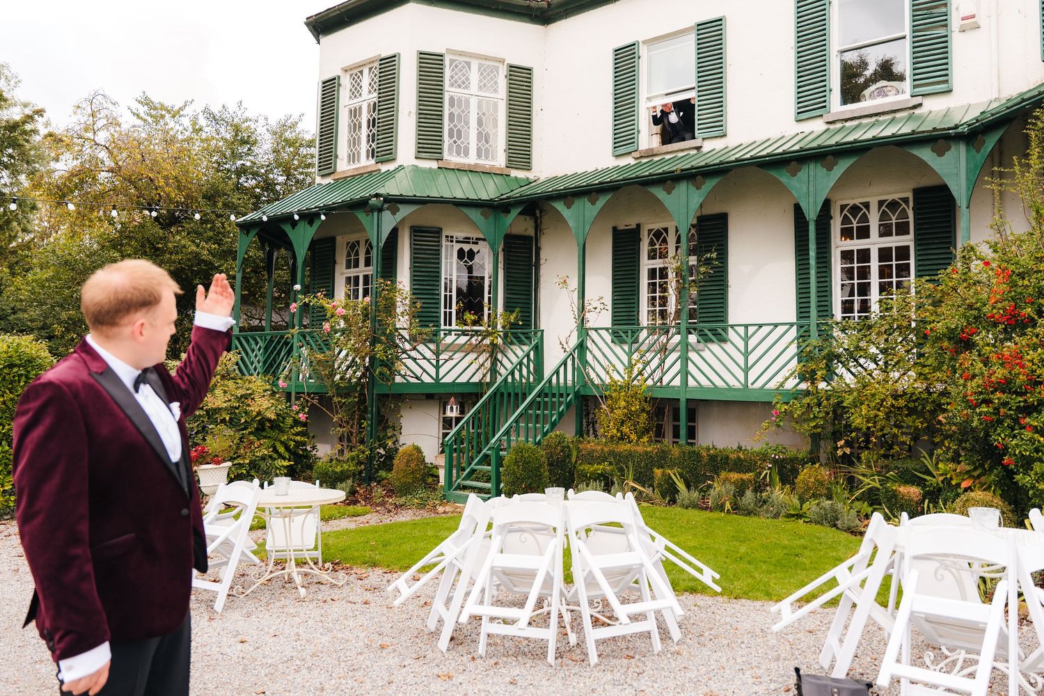 Person in burgundy suit waving at historic green and white house with outdoor seating setup. LGBTQ Ashley Park Wedding Photographer