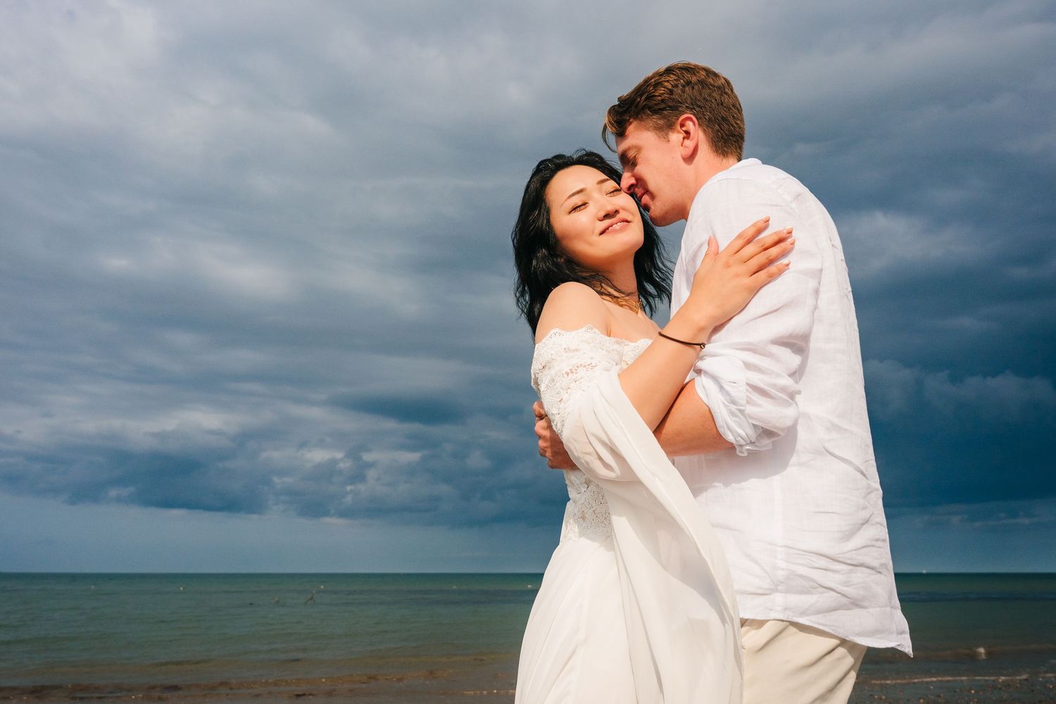 Couple in white embracing and laughing on a beach with dramatic stormy clouds overhead.