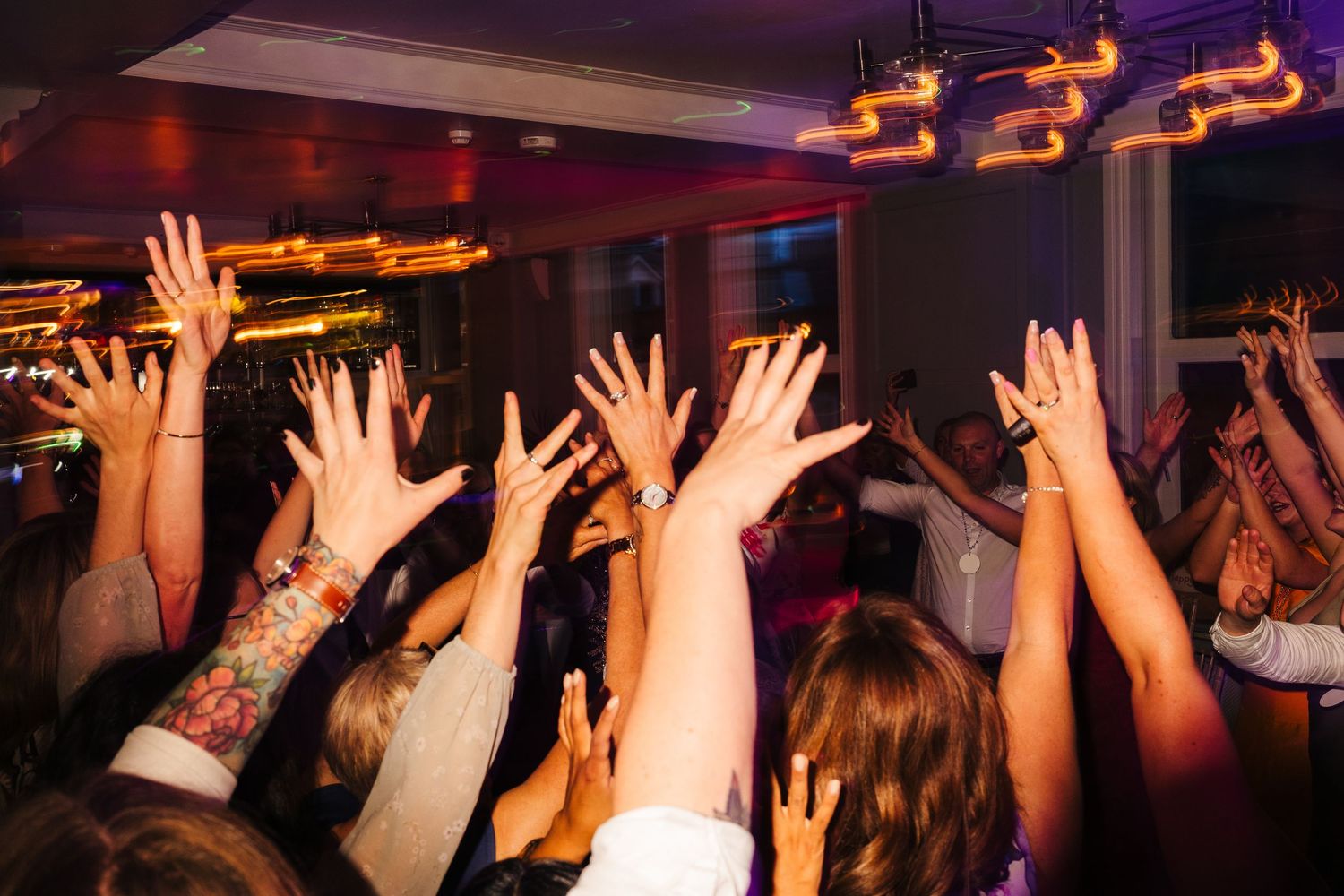 Crowd of wedding guests raise their hands on the dance floor under amber lighting. PHOTOGRAPHER Dublin Ireland Wedding