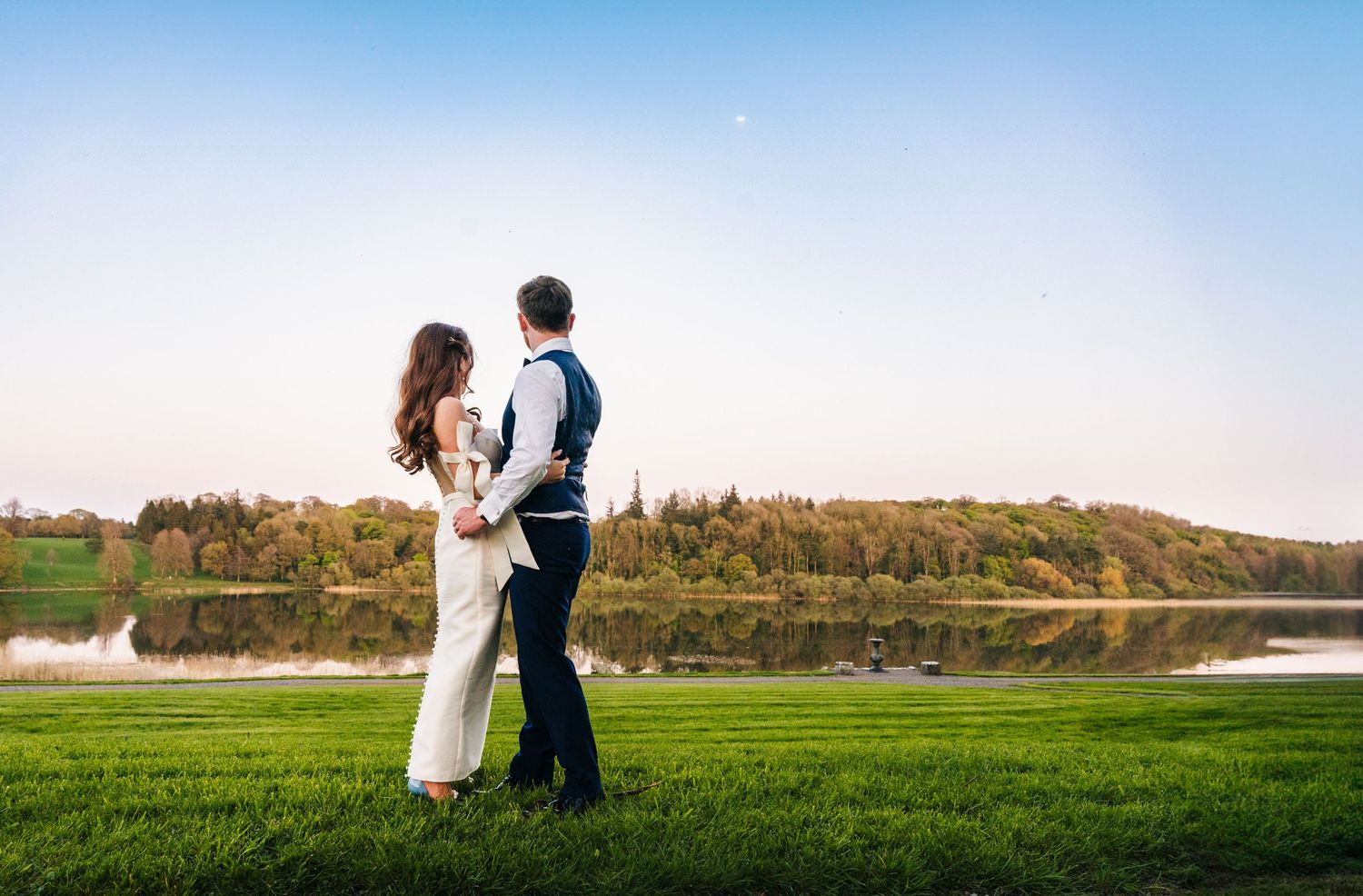 Castle Leslie Wedding Photographer Dublin Romantic sunset portrait of newlyweds standing on green grass with lake and hills in background.