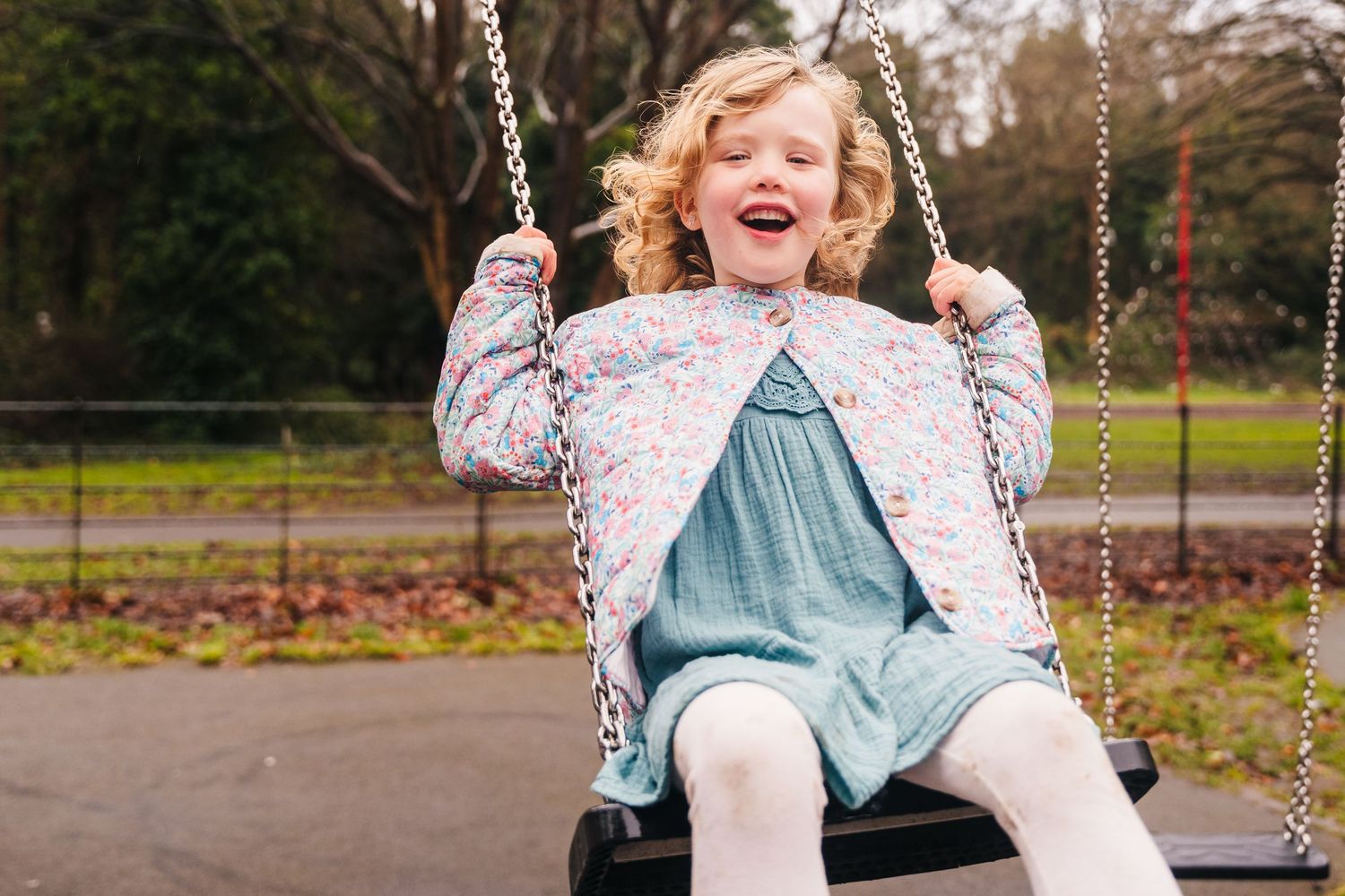 Family photographer Dublin Ireland  A carefree child laughing while swinging at the park, wearing a floral jacket and blue dress.