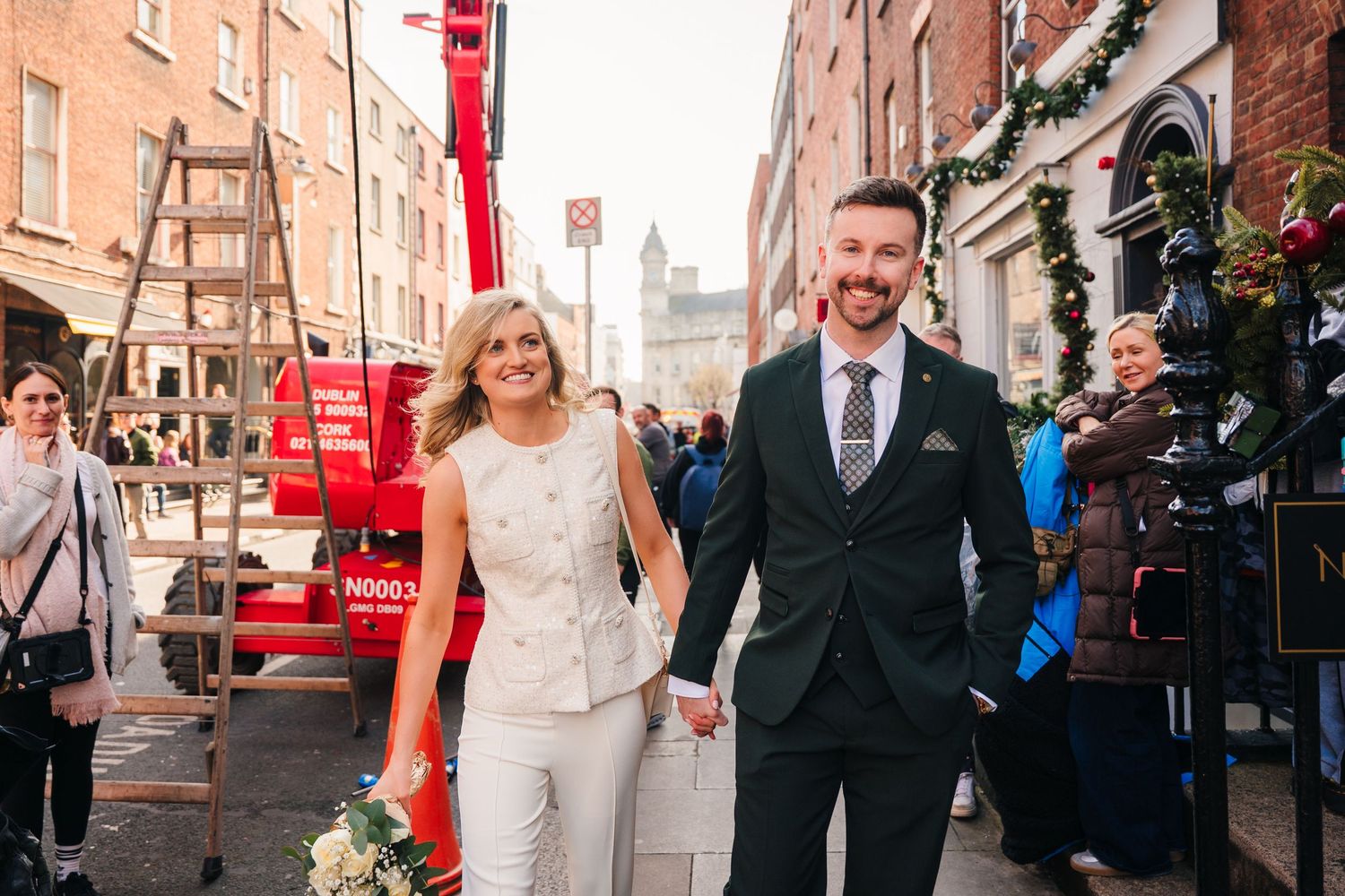 A couple walks down a decorated Dublin street with red banners and historic brick buildings. City Hall Dublin wedding photogrAPher
