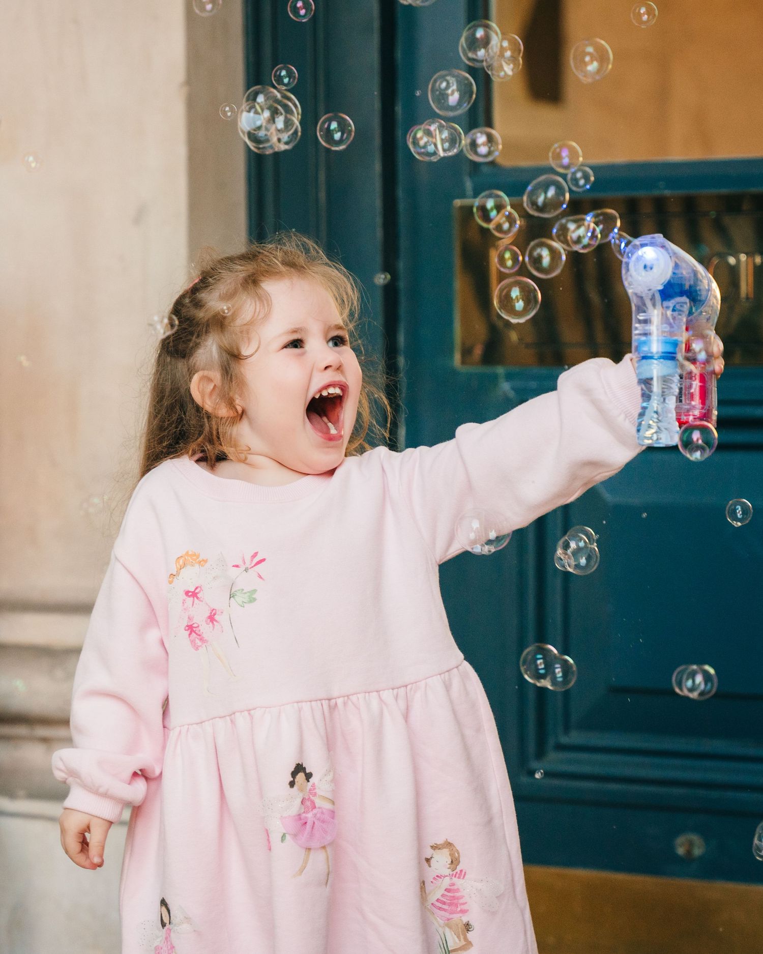A delighted child in a pink dress plays with bubbles against a green door background. City Hall Dublin wedding photogrAPher