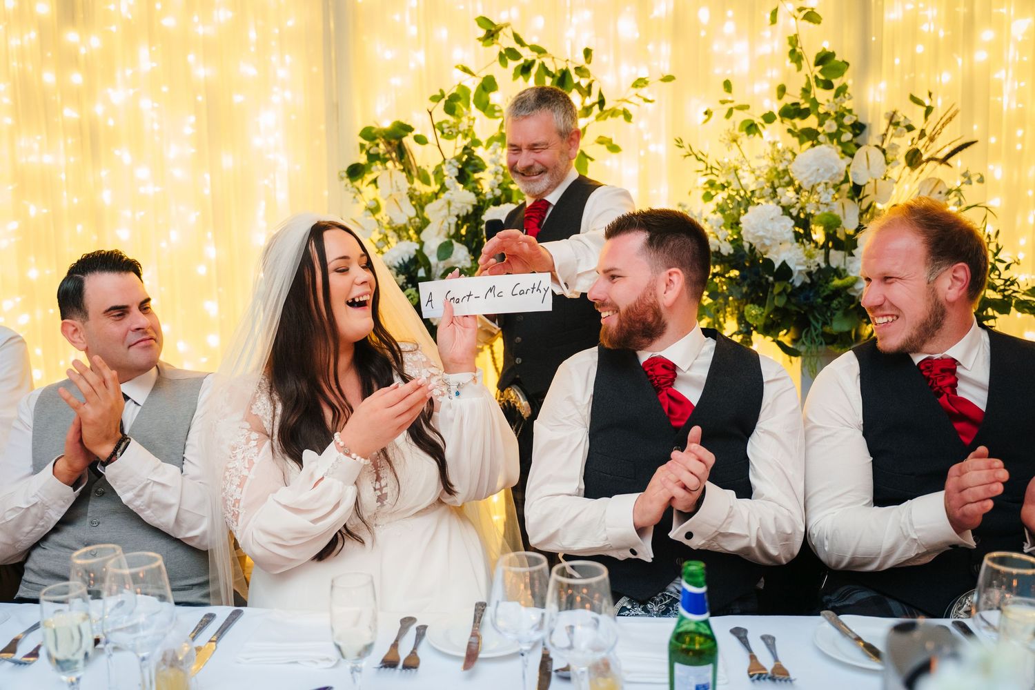 Wedding reception guests laughing together at decorated dinner table with twinkle light backdrop. Portmarnock Hotel Wedding PHOTOGRAPHER Dub