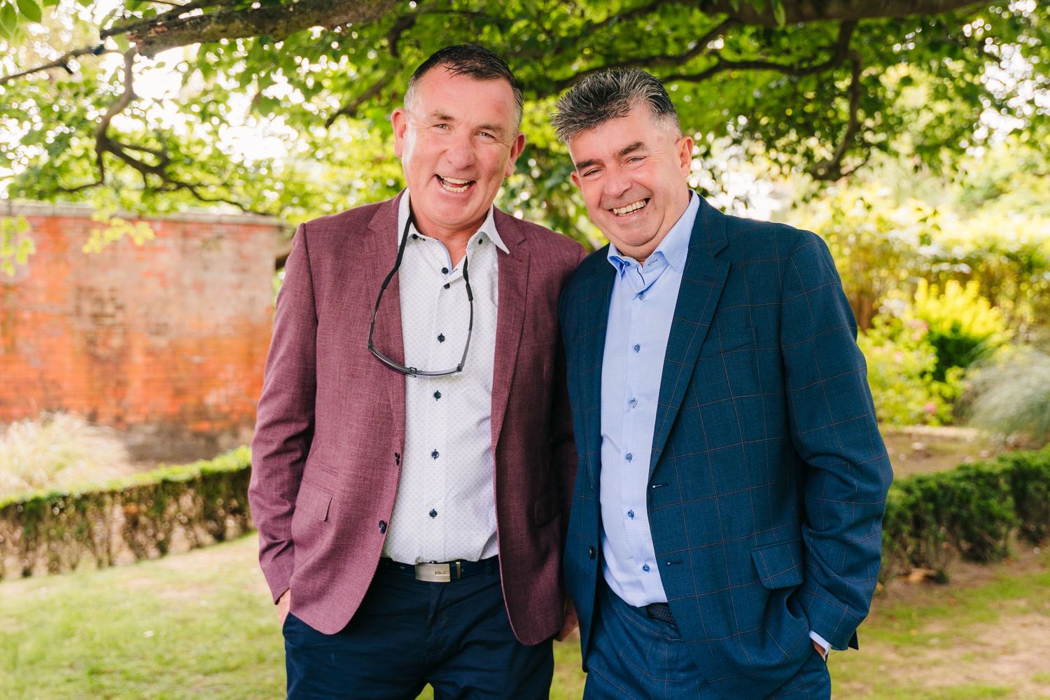 Two smiling friends stand together outdoors wearing dress shirts and blazers - one in mauve and one in teal. Portmarnock Hotel Wedding PHOTO