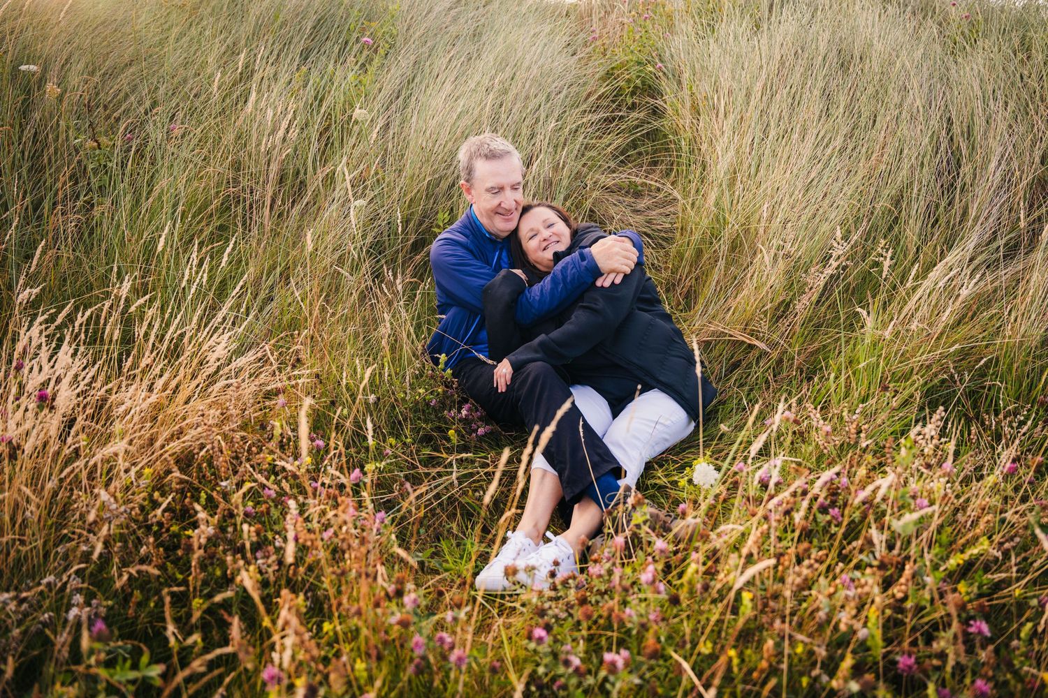 A joyful couple embraces while sitting among tall grass and wildflowers on a beautiful autumn day. PHOTOGRAPHER Dublin ireland