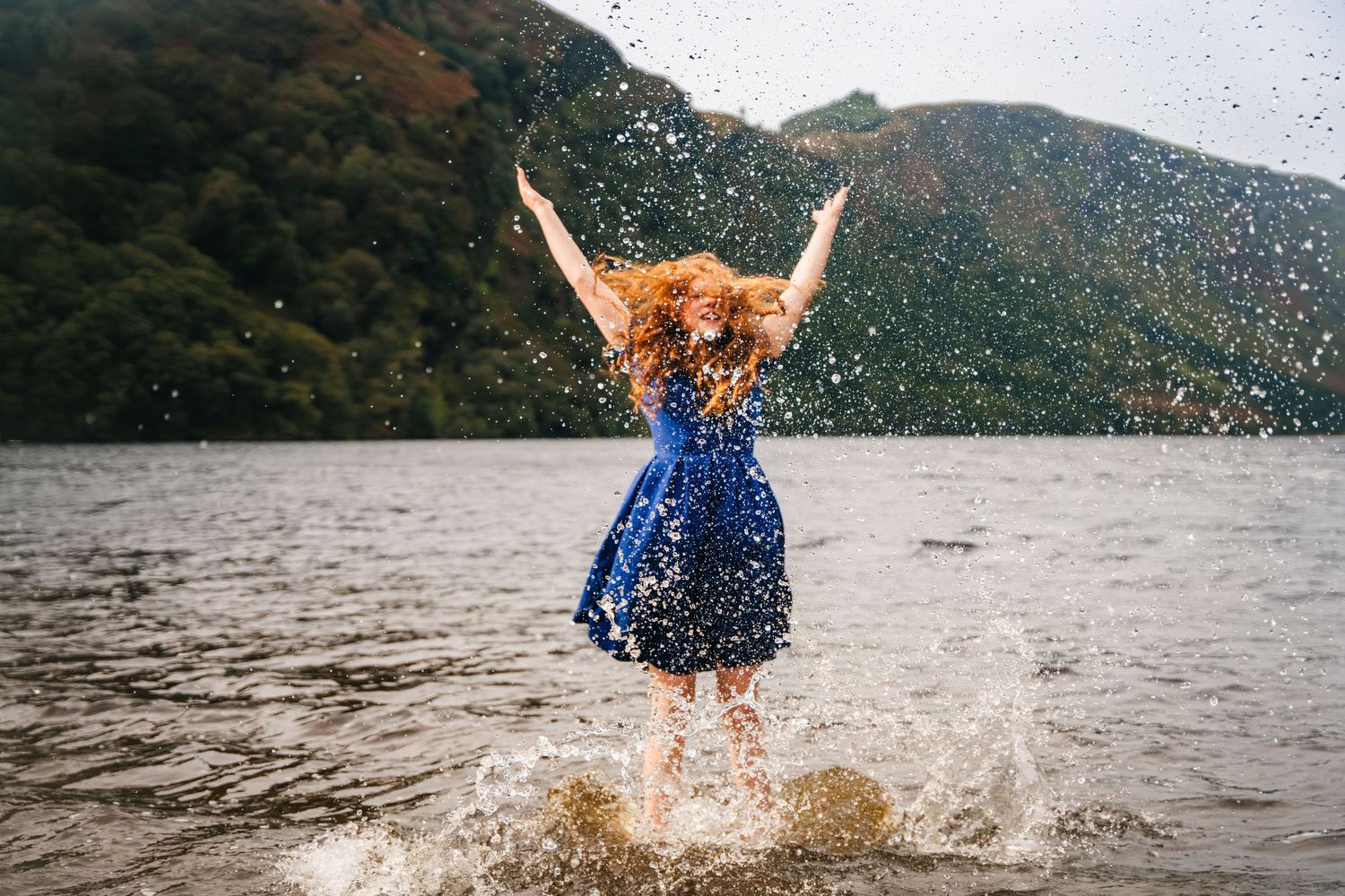 Someone with red hair in a blue dress jumps joyfully into a lake surrounded by mountains, creating a splash. family PHOTOGRAPHER Dublin