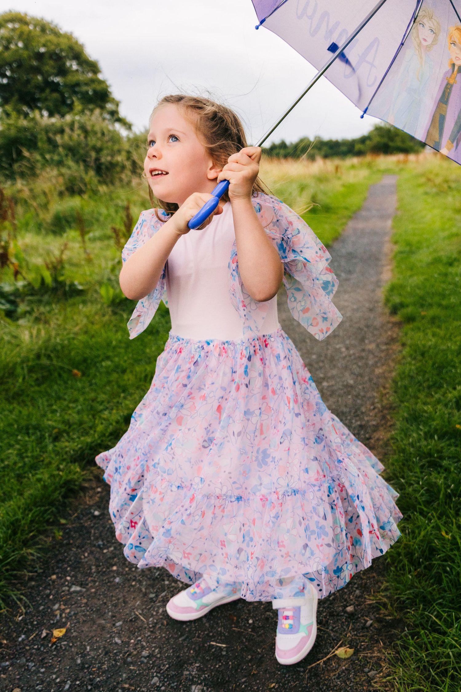 A young child in a floral dress twirls with a purple umbrella on a countryside path during a cloudy day. family PHOTOGRAPHER Dublin