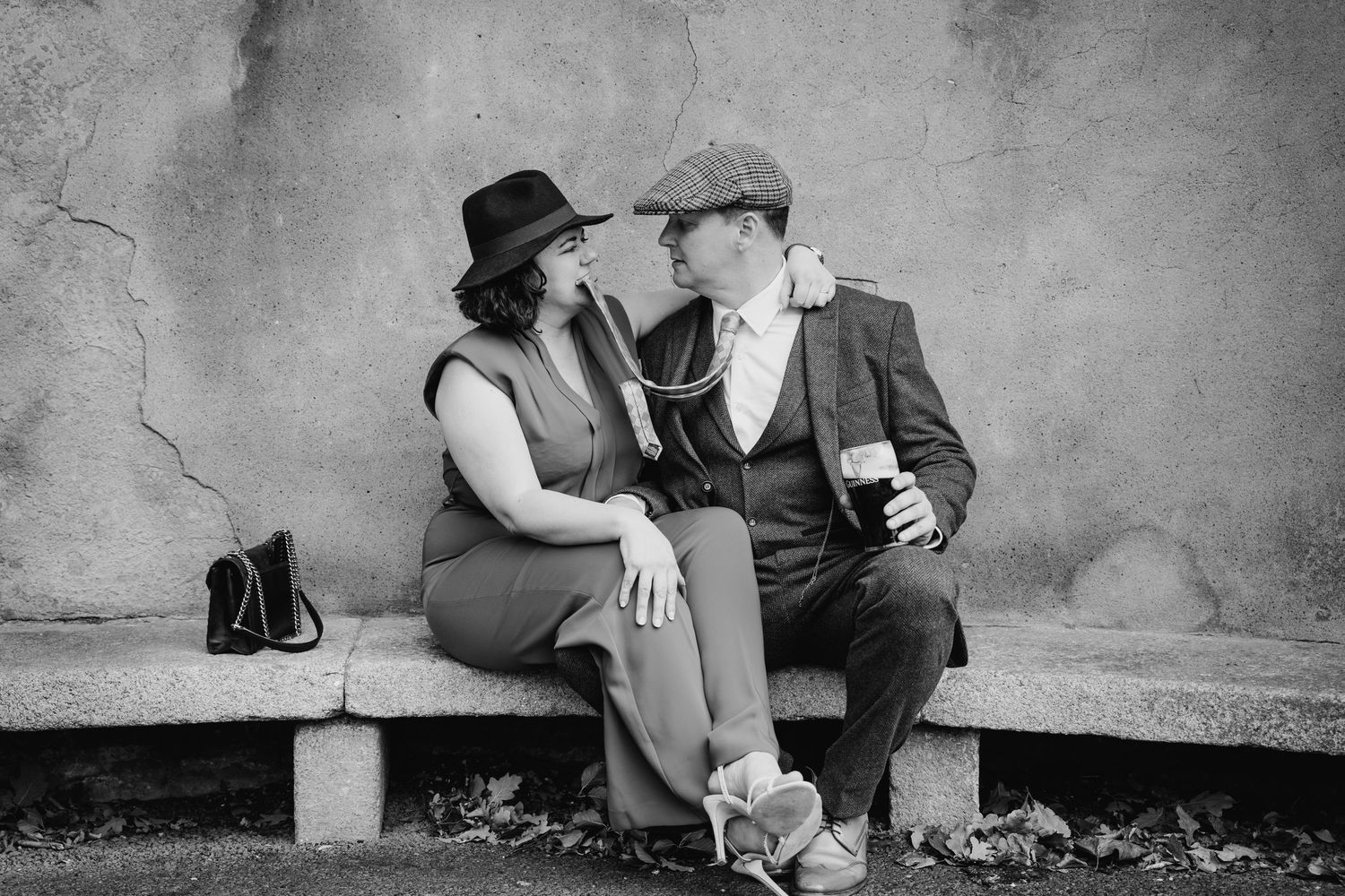 A vintage black and white photograph of a couple in period clothing sitting intimately on a bench against a textured wall. PHOTOGRAPHER Dubl