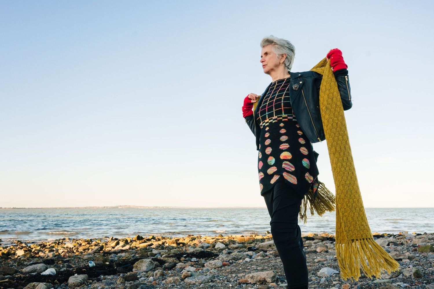 Person in black coat stands on rocky beach holding yellow yarn while ocean waves crash in background. professional headshots Dublin