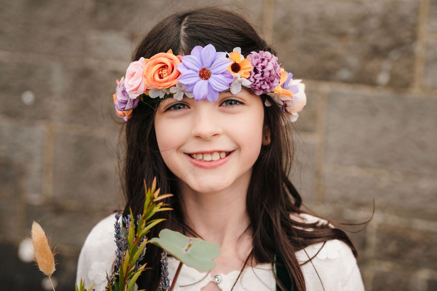 A person wearing a delicate floral crown made of pink and purple flowers smiles against a rustic wooden background.